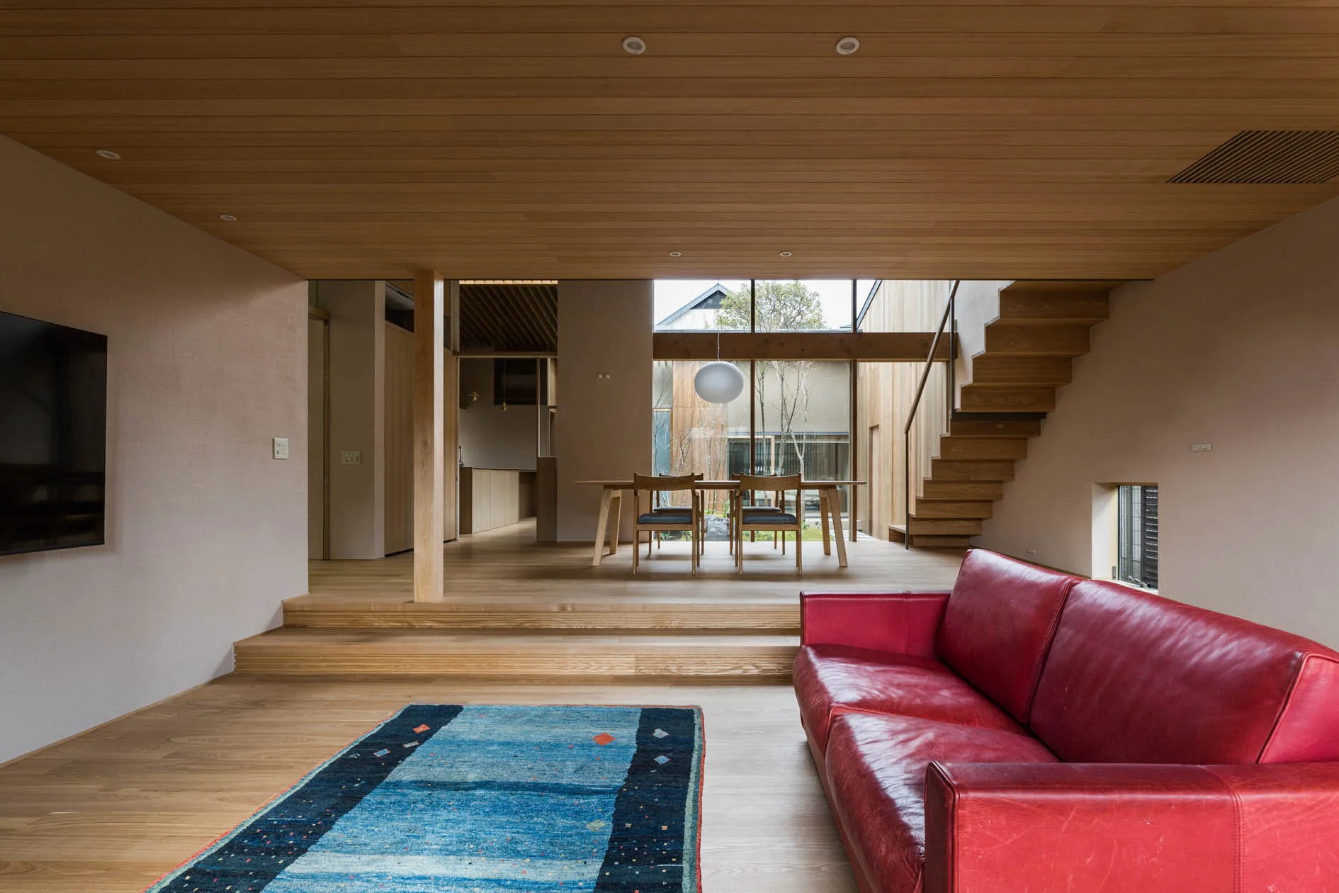 A sunken living room with wood-paneled ceilings, a red leather sofa on a blue rug, and stairs leading to an elevated dining space.