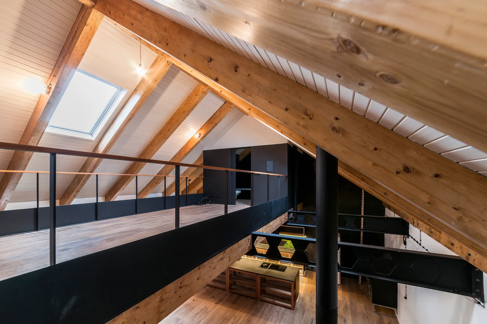 An elevated wood mezzanine overlooks a lower level kitchen under a vaulted ceiling with exposed timber rafters and a skylight.