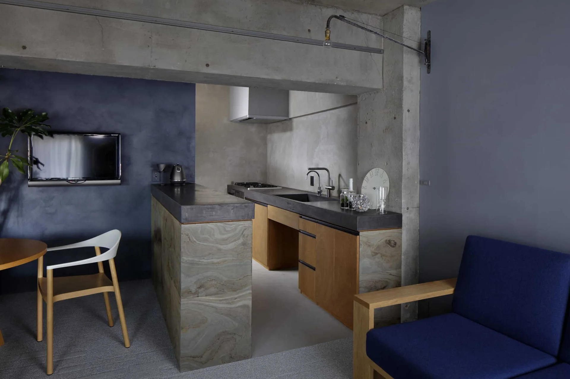 A modern kitchen features an exposed concrete beam above a swirling stone-clad island, wood cabinetry, and a dark blue accent wall.
