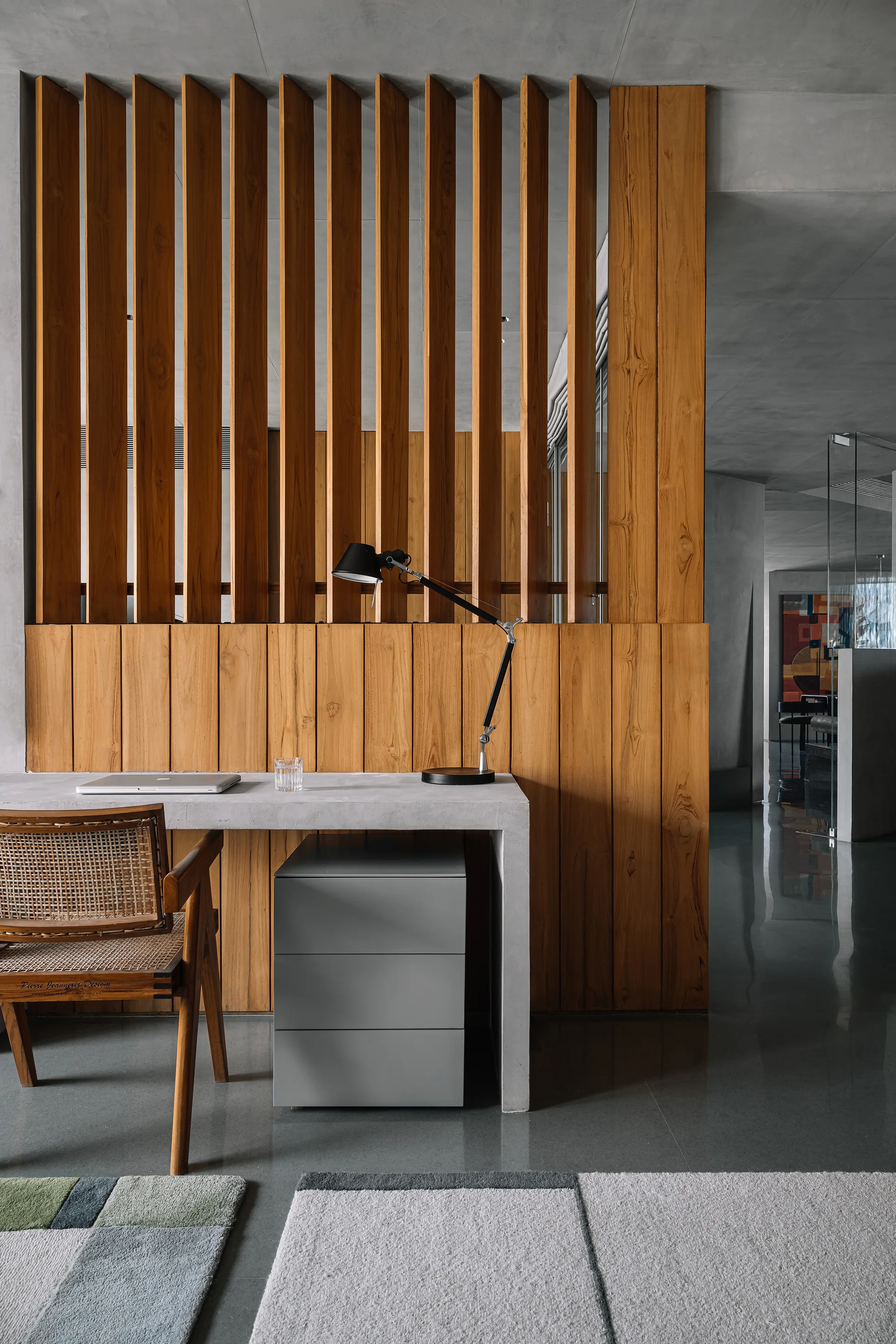 A minimalist home office workspace features a concrete desk with a black desk lamp, a rattan chair, and a vertical wooden slat partition wall.