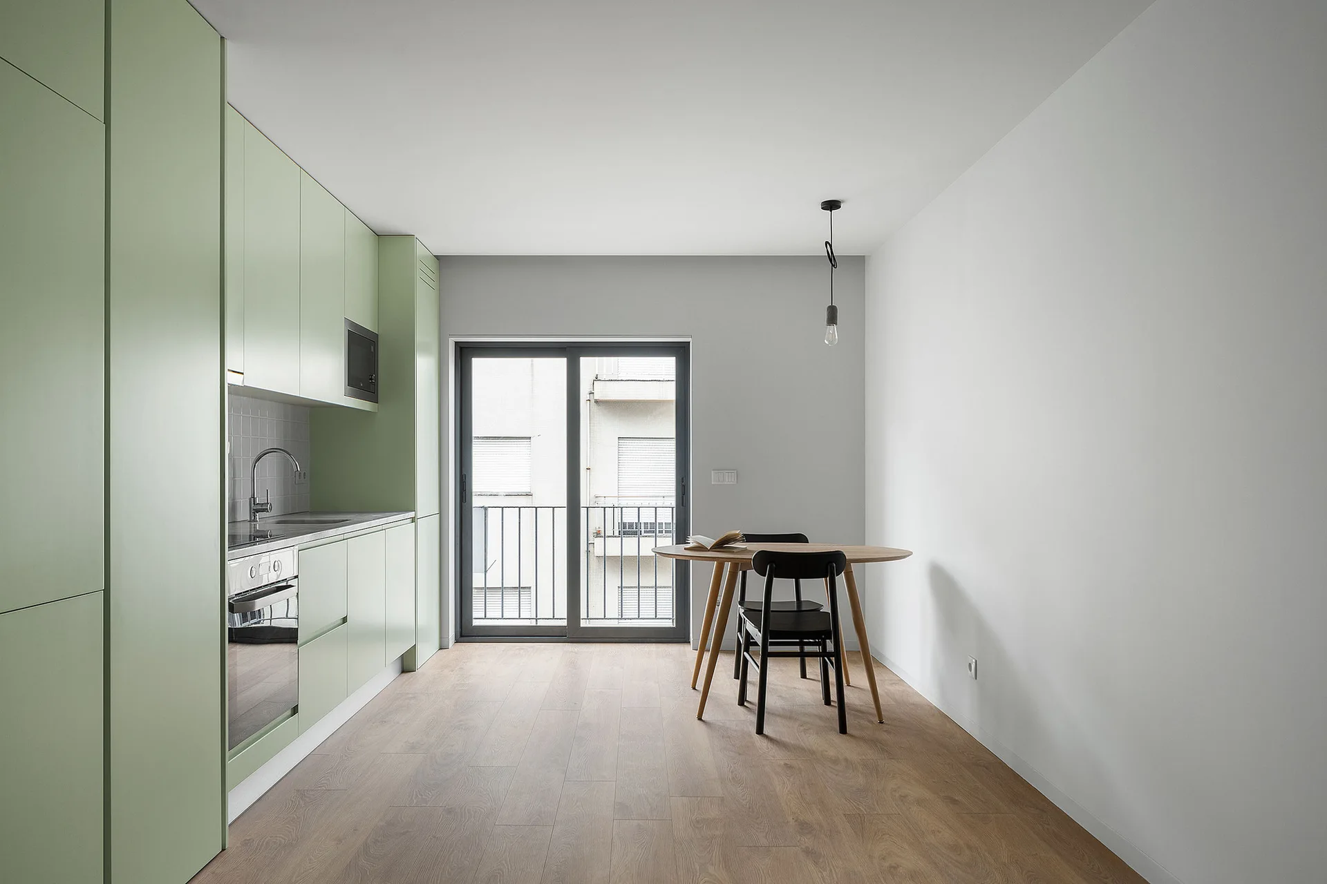A minimalist kitchen features floor-to-ceiling pale green cabinets, a round wooden dining table with black chairs, and light wood flooring.