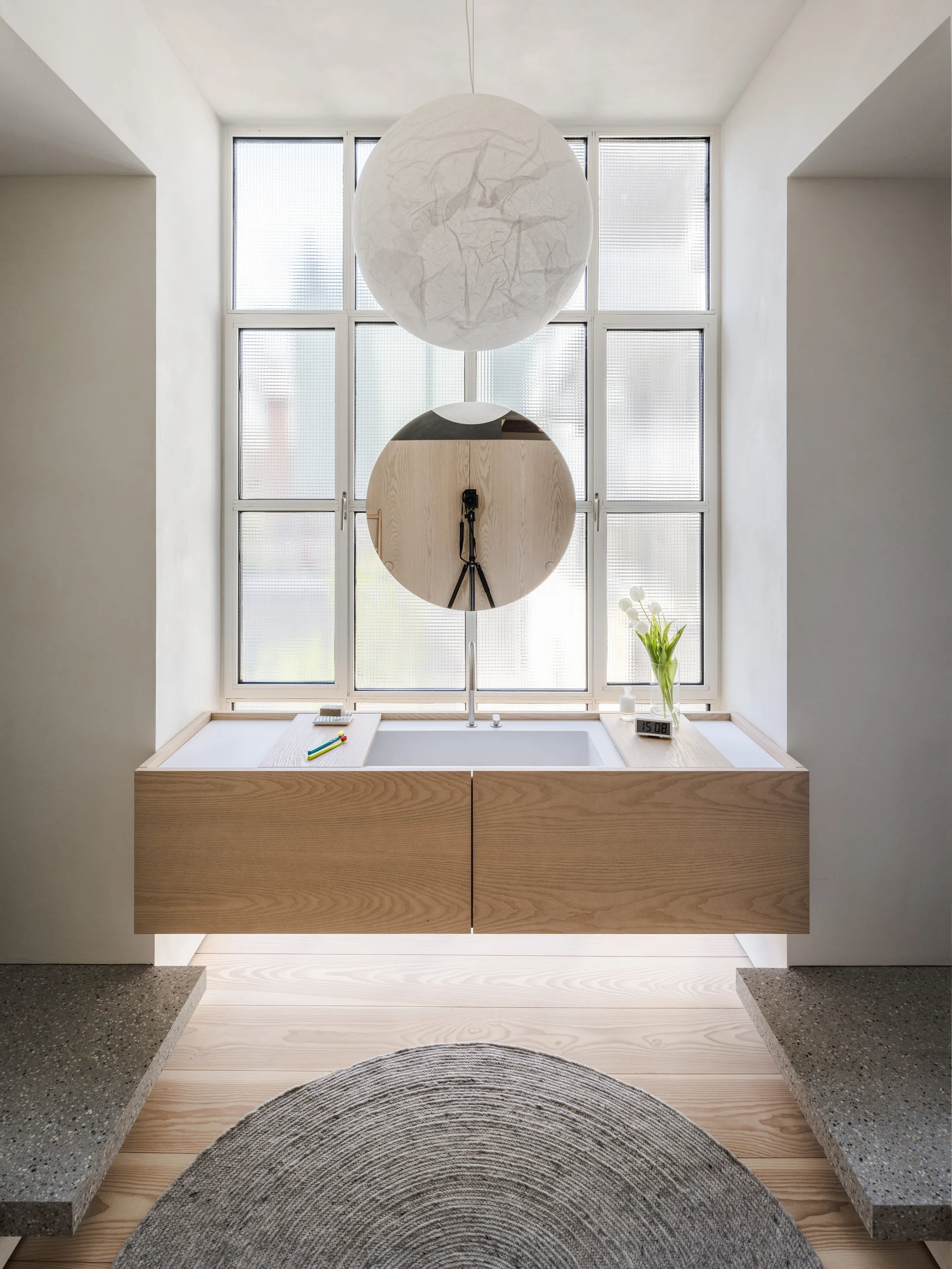 A minimalist bathroom features a floating wood vanity with an integrated sink set in front of a tall gridded window and a round mirror.