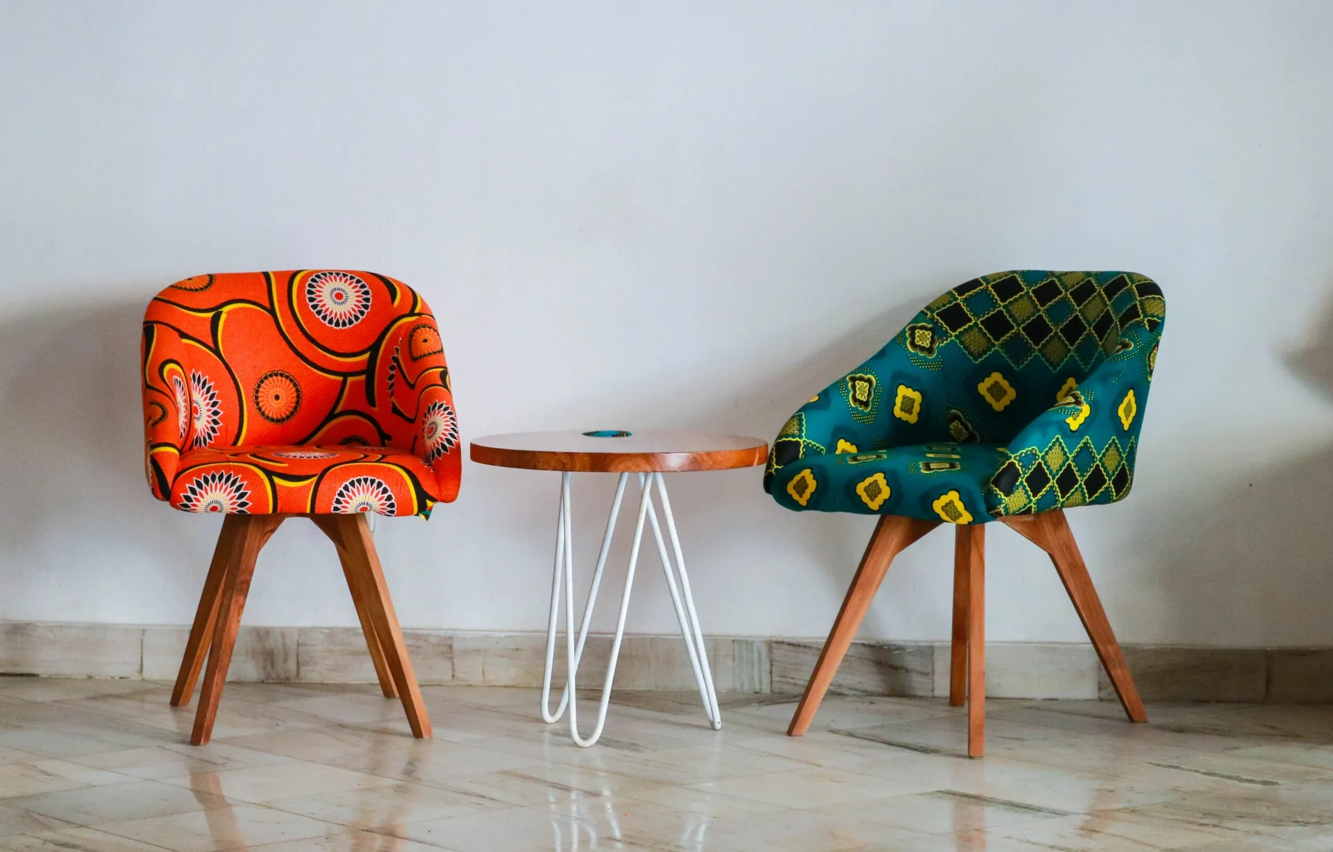 Two upholstered armchairs with orange and teal patterned fabric sit on either side of a round wooden table with white hairpin legs.