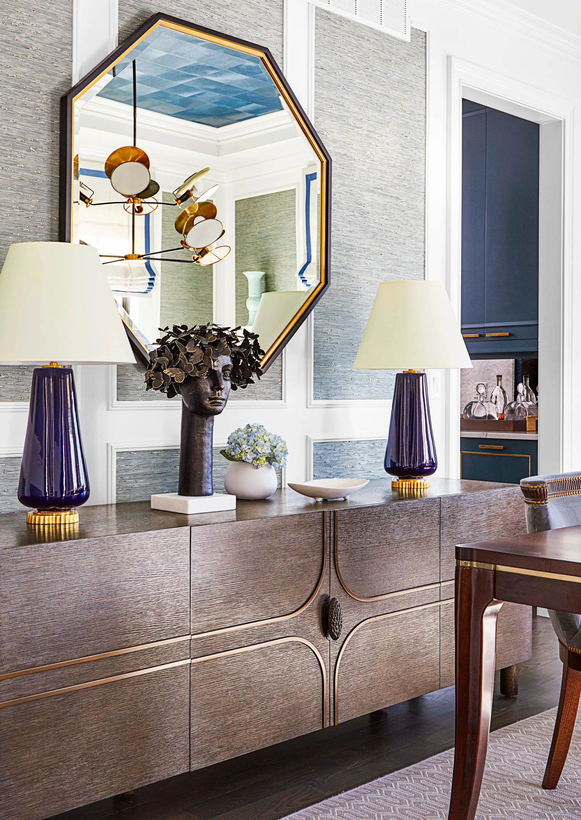 A wooden sideboard features two glossy navy lamps, a bronze bust sculpture, and an octagonal mirror against paneled walls with textured wallpaper.