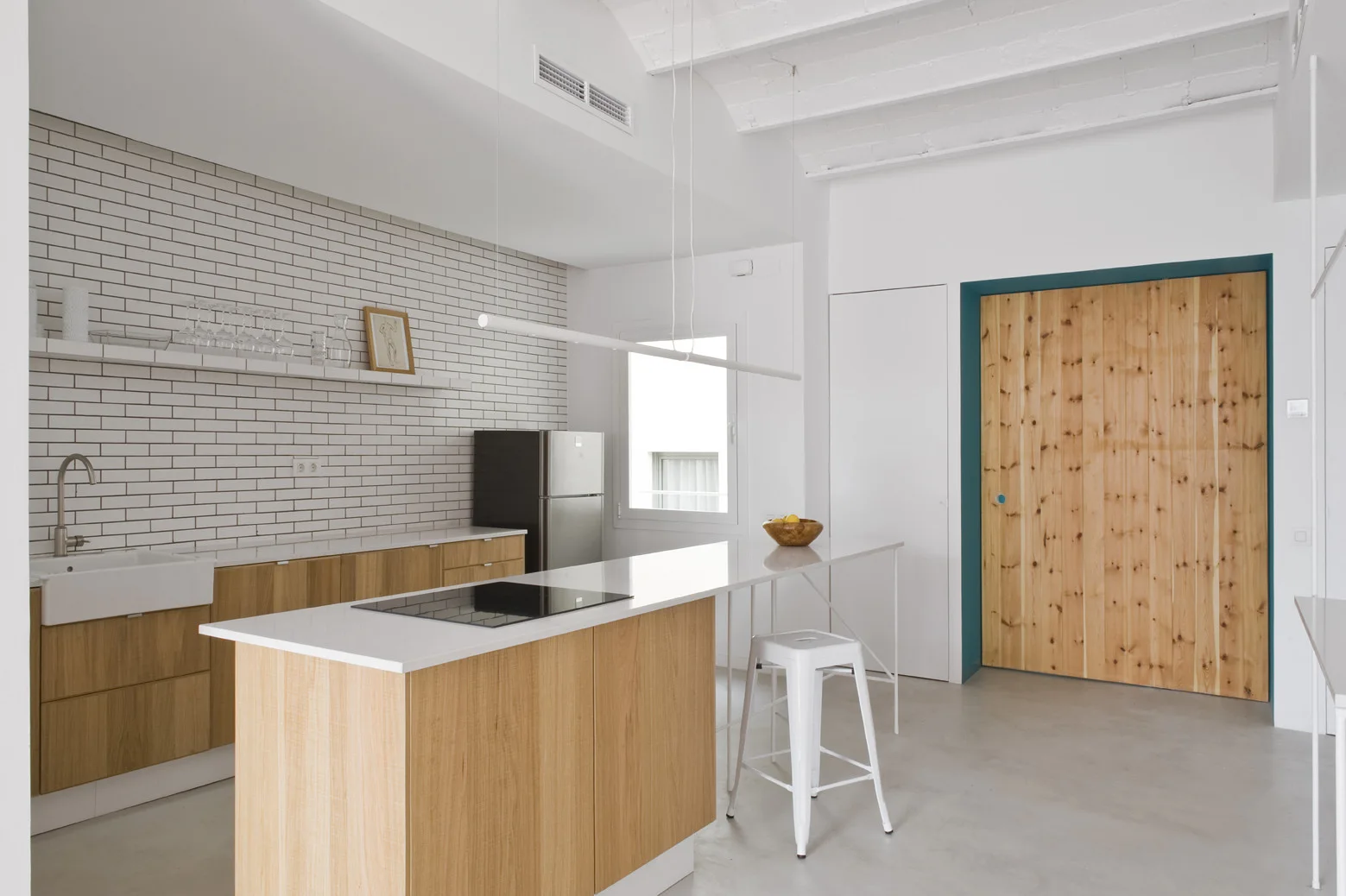 A minimalist kitchen features wood cabinets, a white brick tile backsplash, a white island with a metal stool, and a large wooden sliding door.