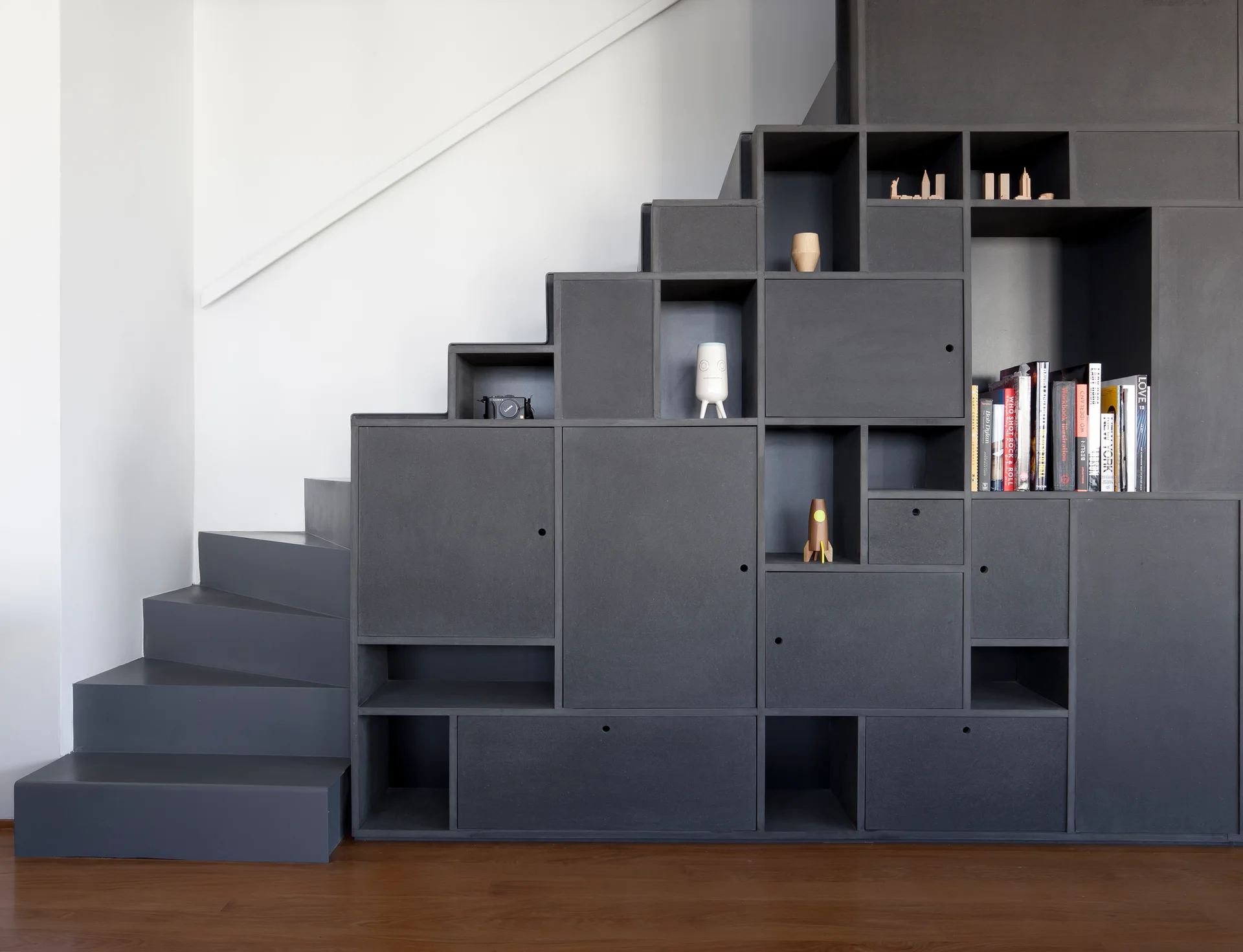A dark grey staircase with integrated storage cabinets, open shelves, and cubbies stands against a white wall on wood flooring.