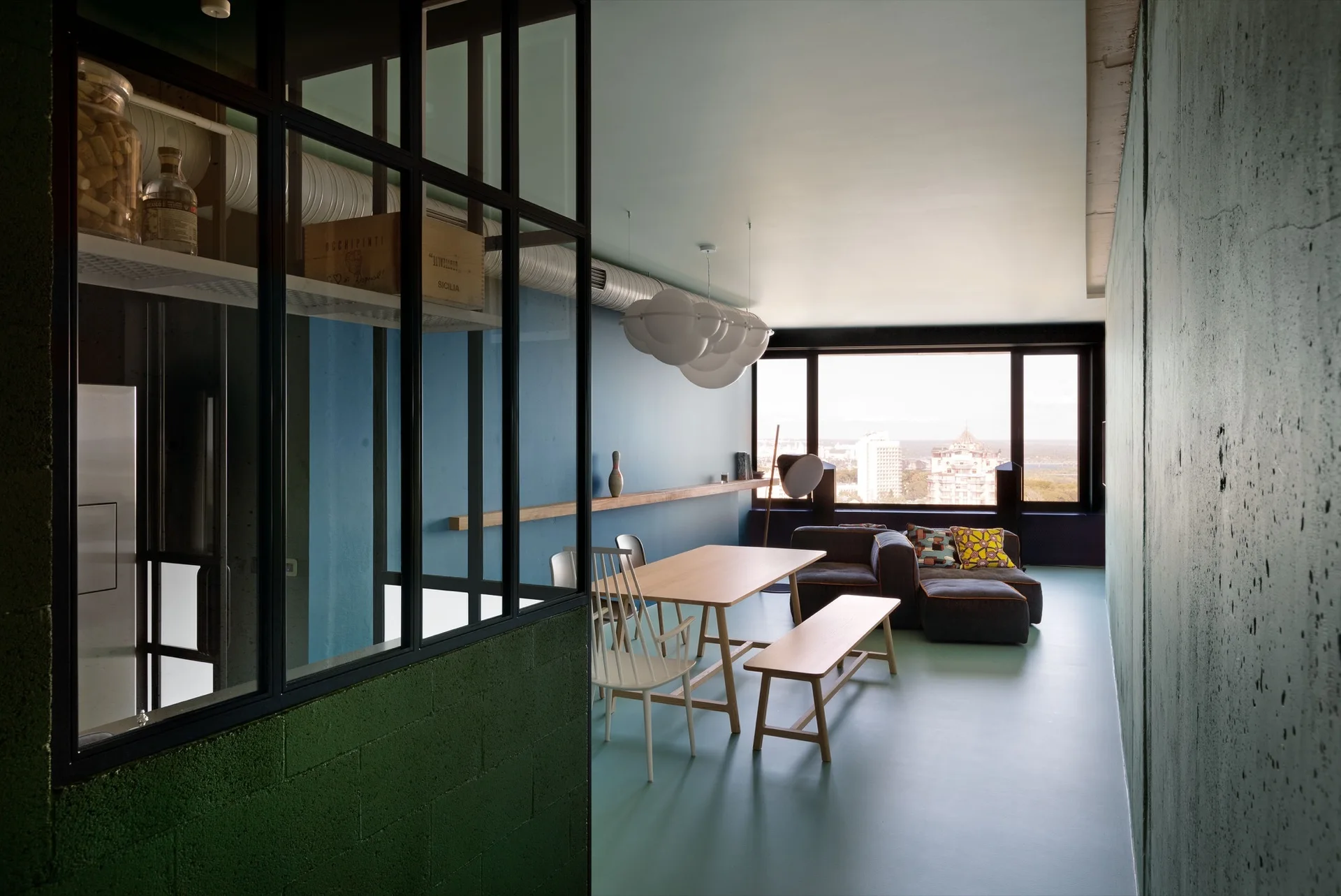 An open-plan apartment interior features a dining table, a low sofa, bold blue and green walls, and a black-framed glass partition.
