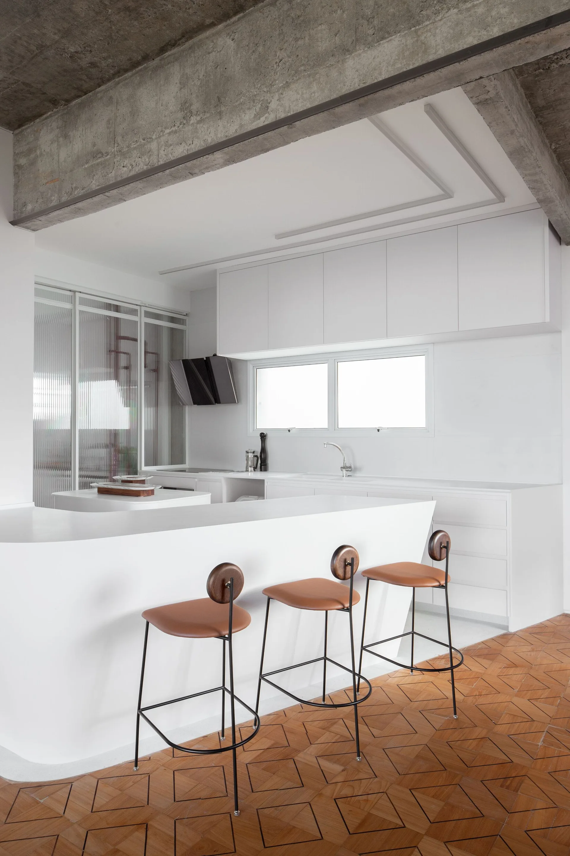 Three leather-seated bar stools stand against a curved white kitchen island under an exposed raw concrete ceiling and beside a ribbed glass divider.
