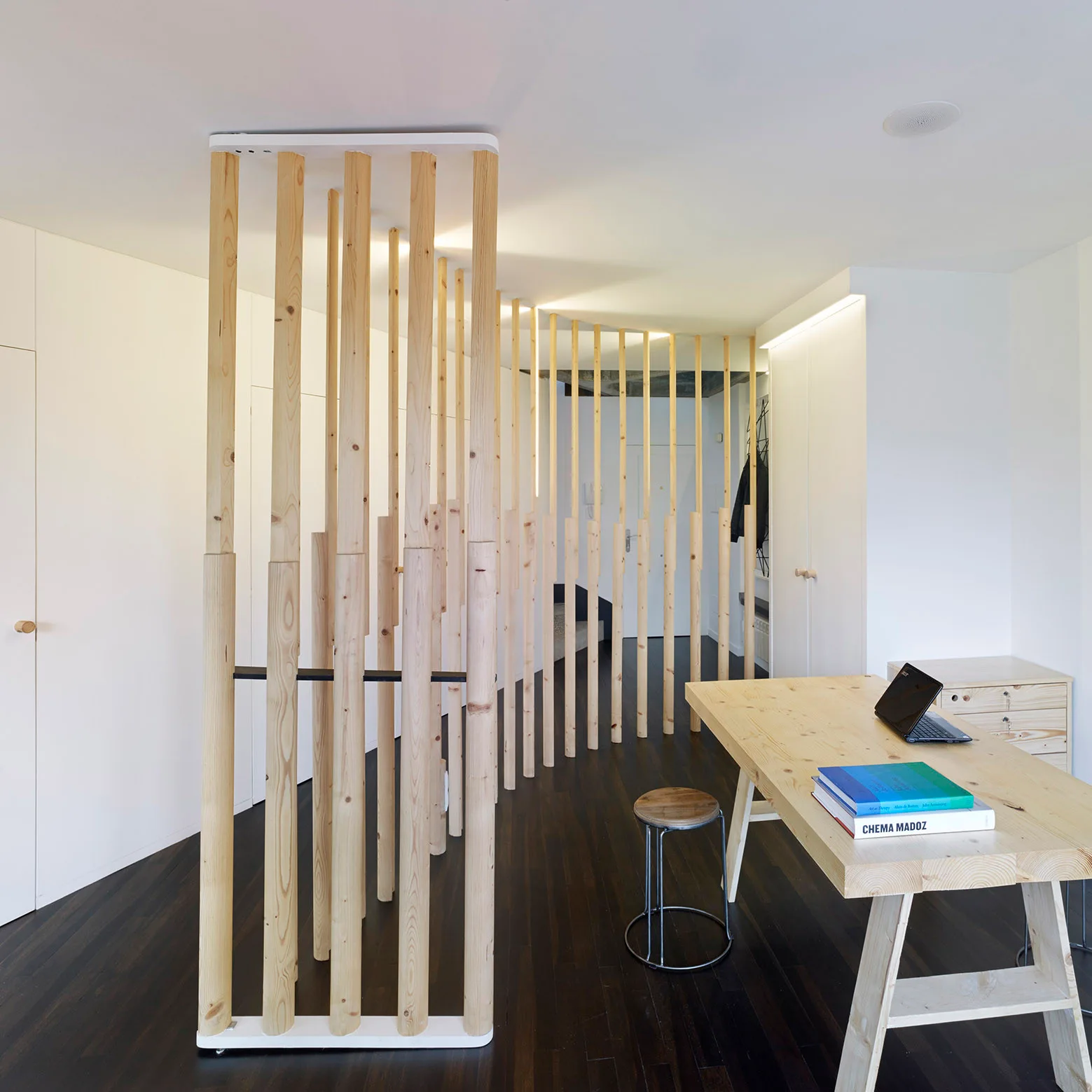 A minimalist home office features a natural wood desk, a small stool, dark wood floors, and a curved room divider made of vertical wooden poles.
