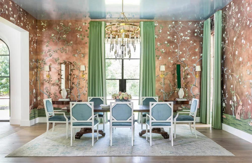 A formal dining room featuring copper floral wallpaper, light blue upholstered chairs, green curtains, and a high-gloss blue ceiling with a brass chandelier.
