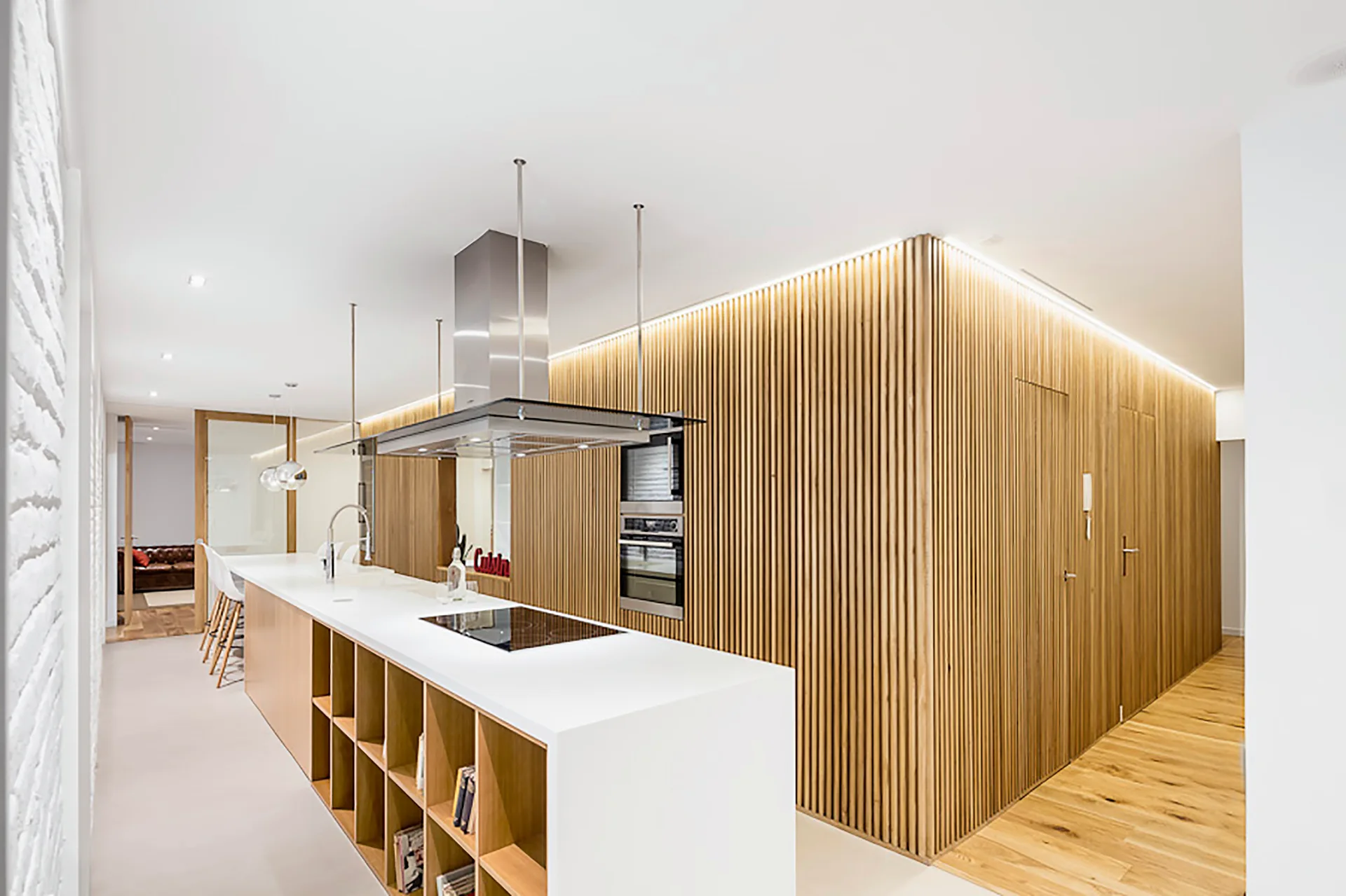 A modern kitchen with a long white island, open shelving, and a feature wall covered in vertical wooden slats with integrated LED lighting.