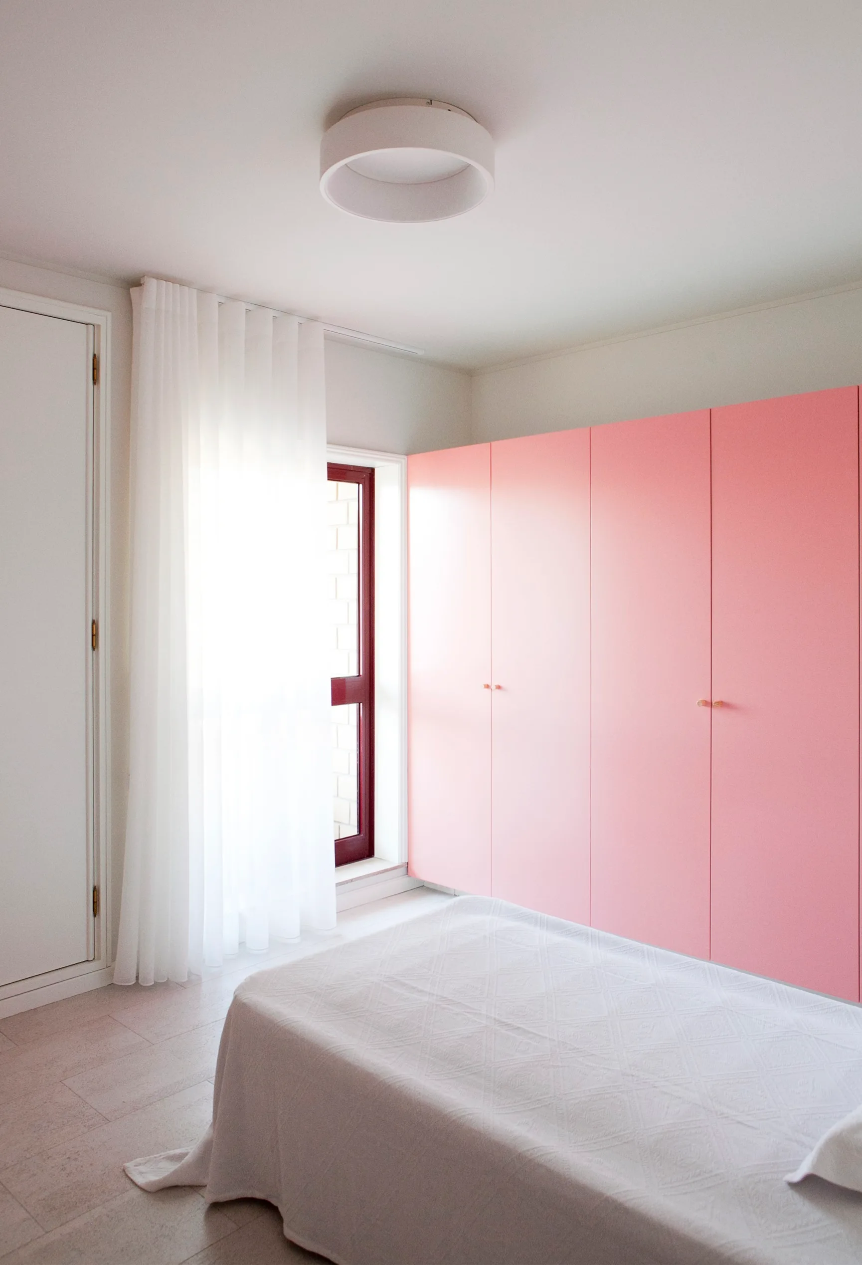 A minimalist bedroom features a large matte pink built-in wardrobe alongside a white bed, sheer curtain, and circular ceiling light.