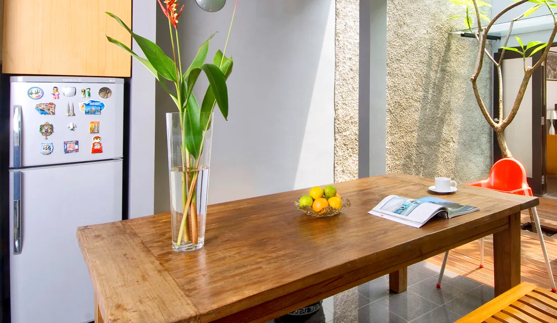 A rustic wooden dining table styled with a tall glass vase sits in an indoor-outdoor dining area next to a textured accent wall.