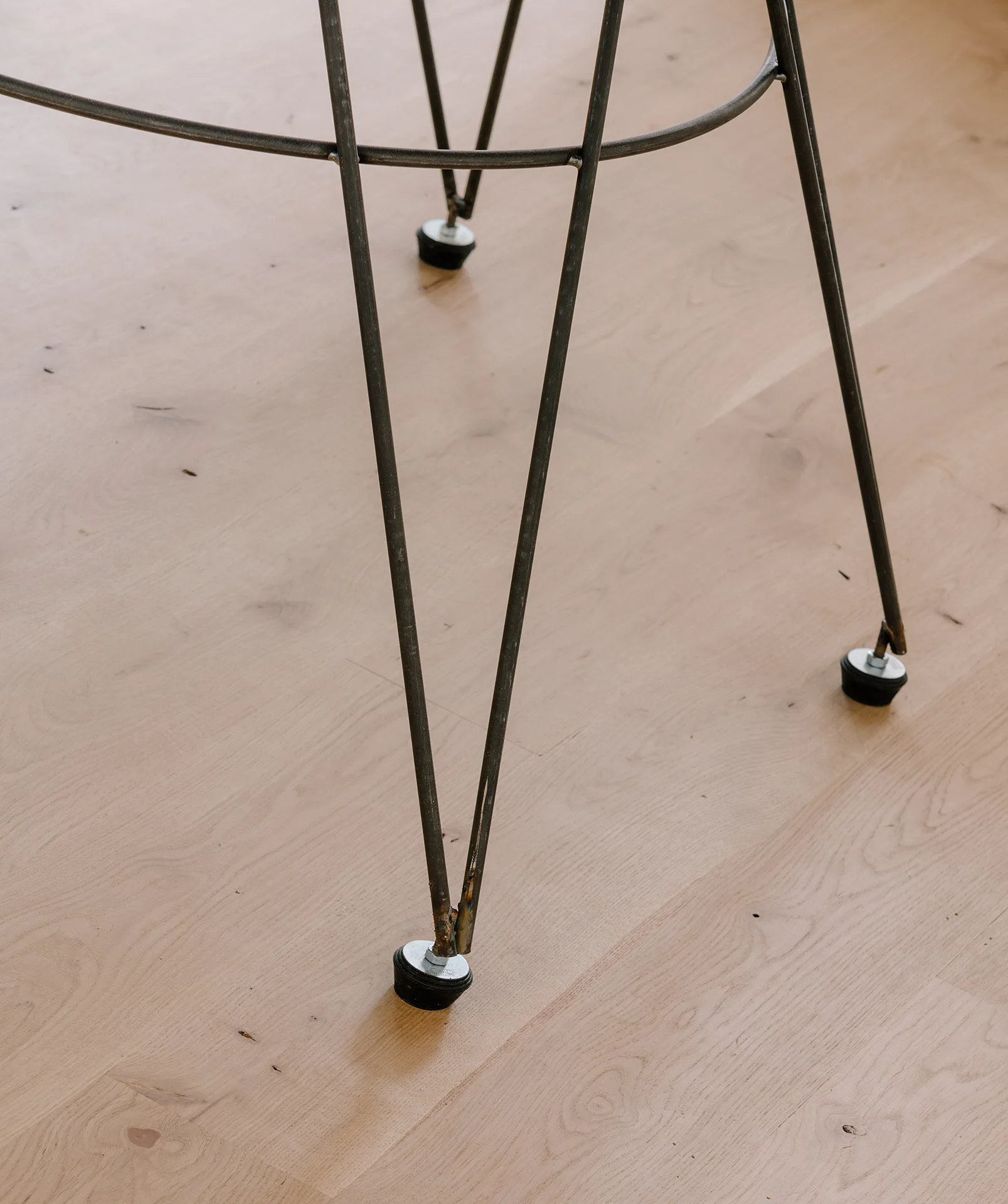 Close-up of thin dark steel hairpin furniture legs with adjustable black rubber protective tips on a light oak wood plank floor.