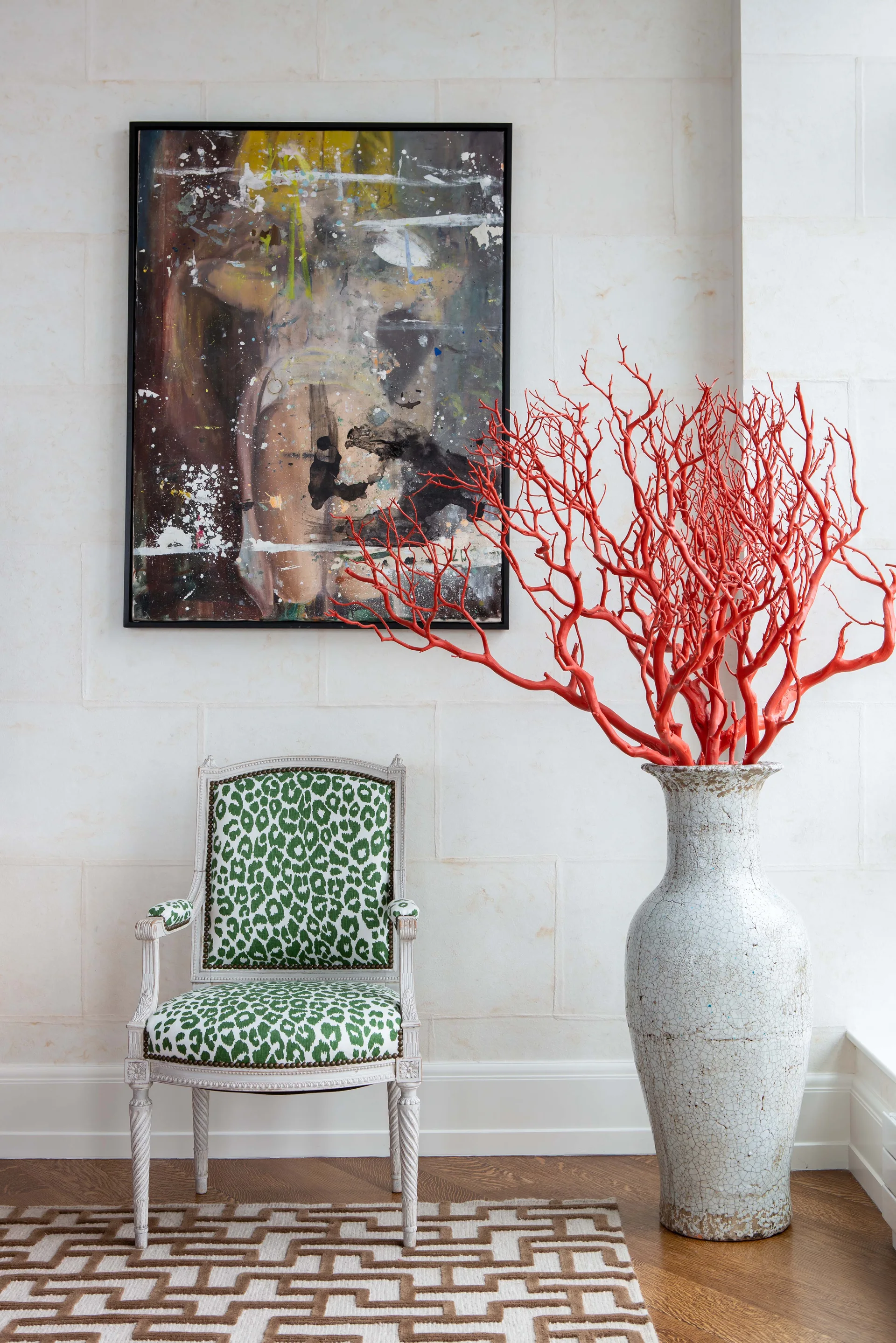 A green leopard-print armchair stands next to a tall white ceramic vase holding bright red branches under an abstract painting on a stone tile wall.