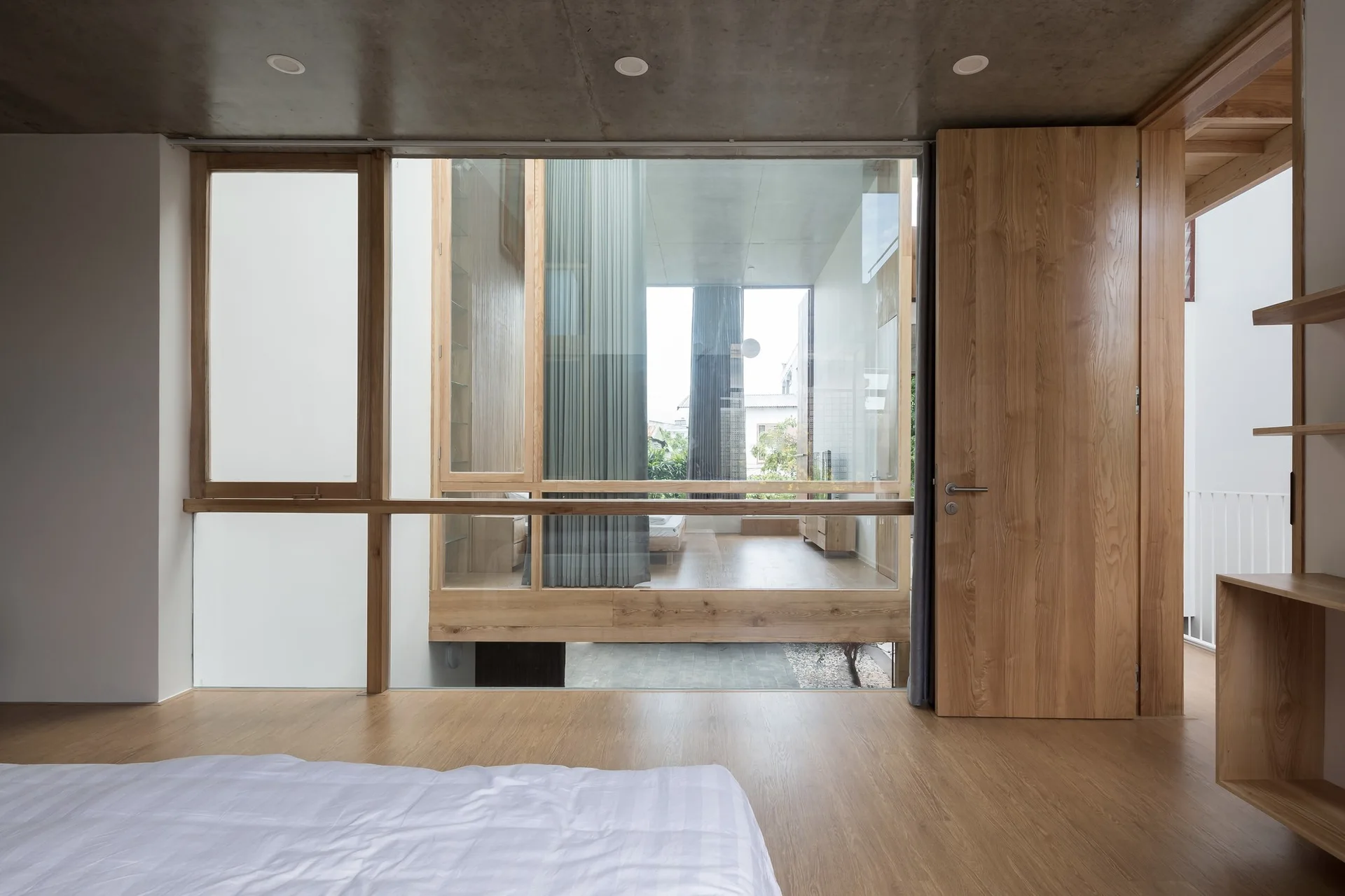 A minimalist bedroom featuring light wood flooring, an exposed concrete ceiling, a matching wood door, and a glass partition looking into an adjacent room.