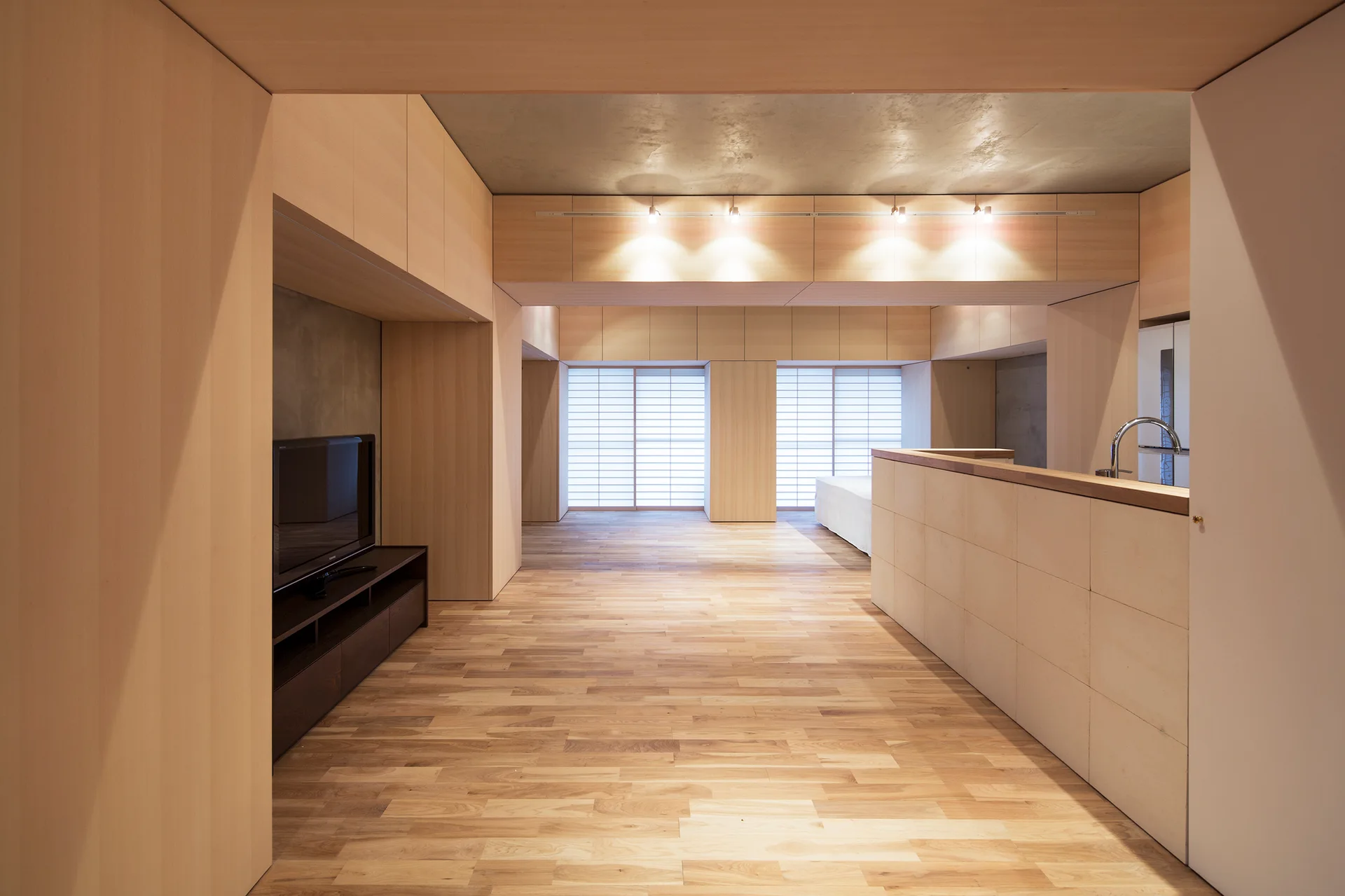 An open-plan apartment featuring extensive light wood wall panels, a polished wood floor, a kitchen counter, and an exposed concrete ceiling.