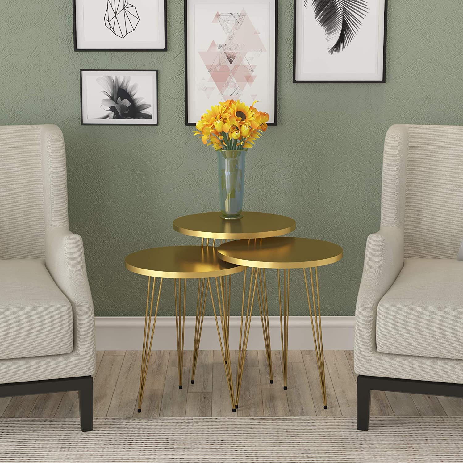 Three gold nesting tables with hairpin legs stand between two beige armchairs against an olive green wall with framed art.