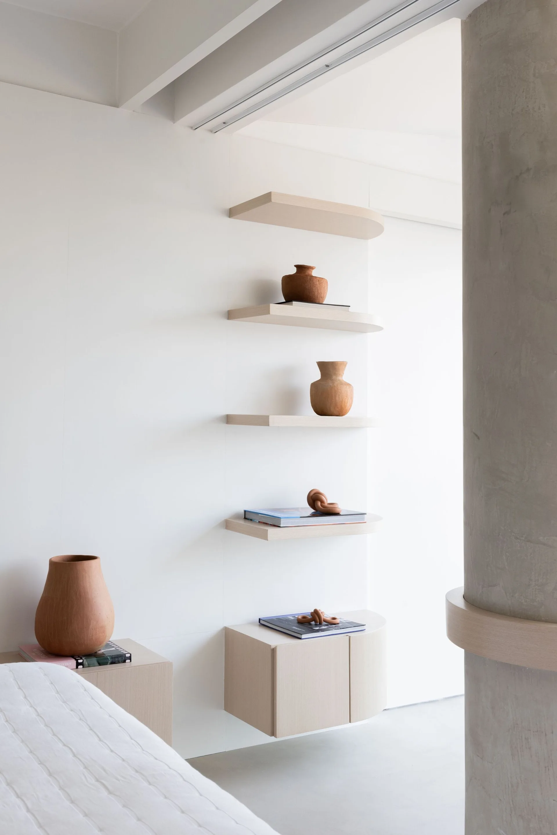 Minimalist light wood floating shelves with curved ends are mounted on a white wall next to a grey concrete column.