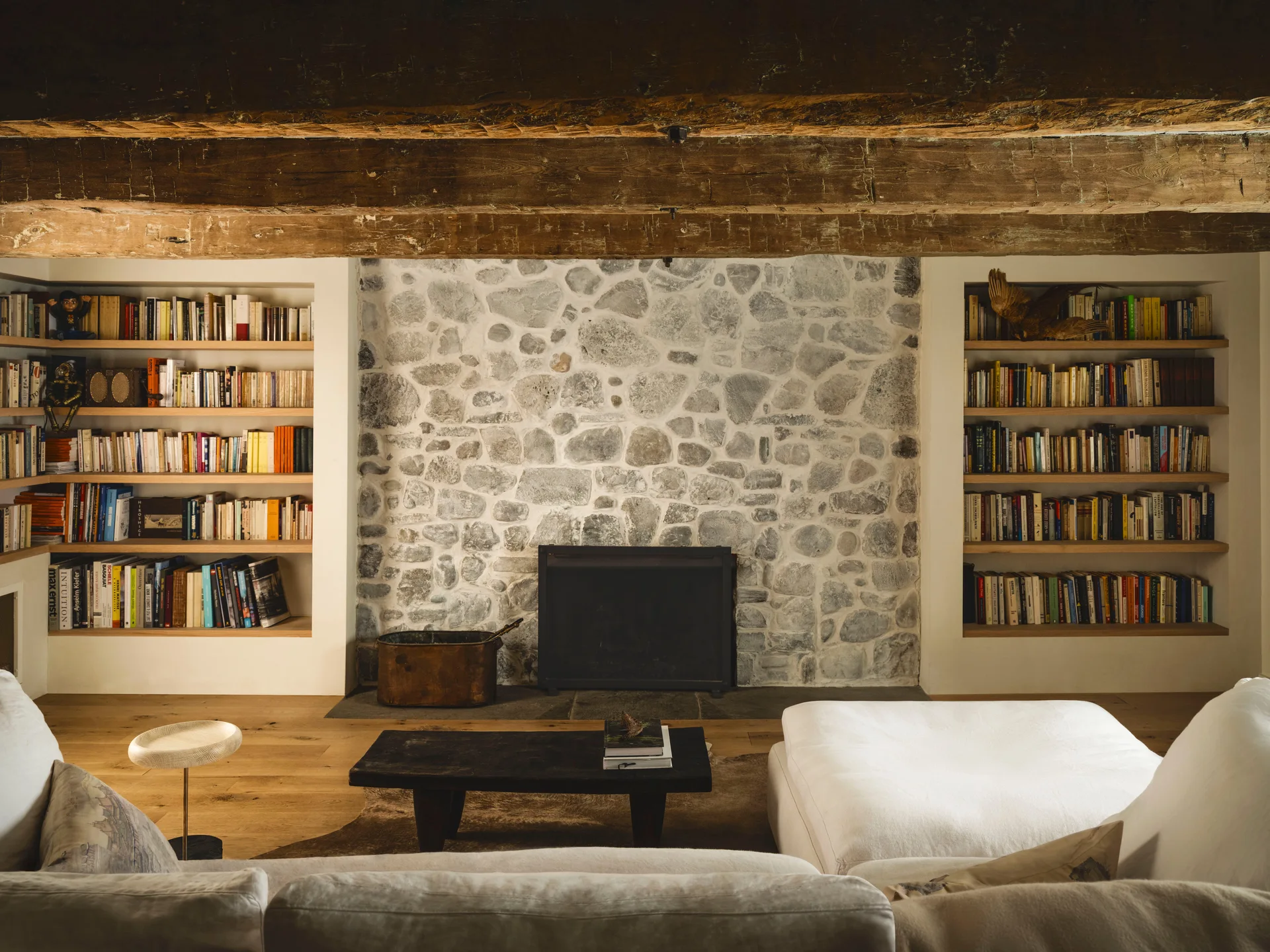 A rustic living room features a textured stone fireplace flanked by built-in wooden bookshelves, beneath massive weathered wood ceiling beams.