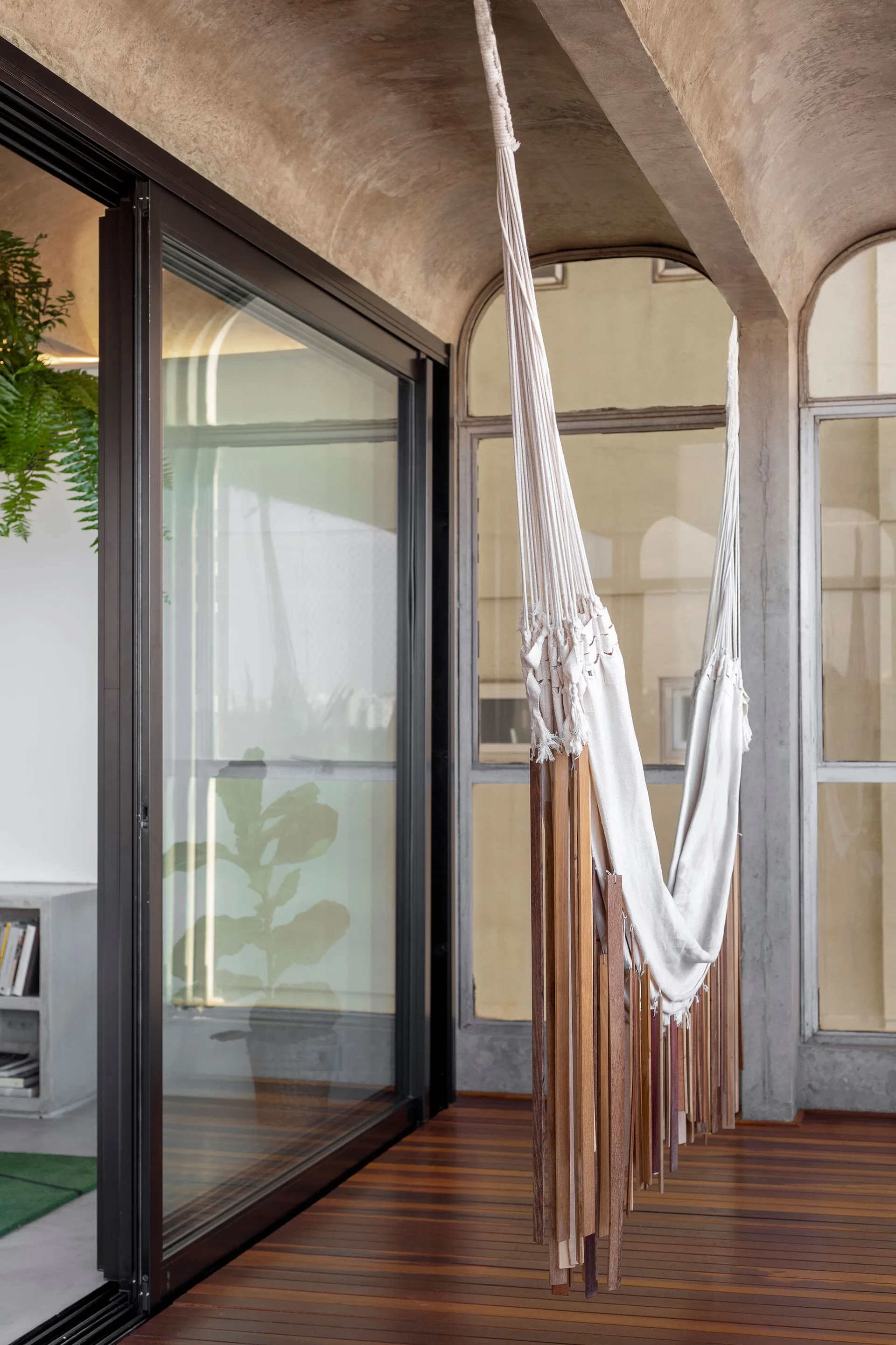 A white fabric hammock hangs on a wooden deck balcony featuring a vaulted concrete ceiling and sliding glass doors.