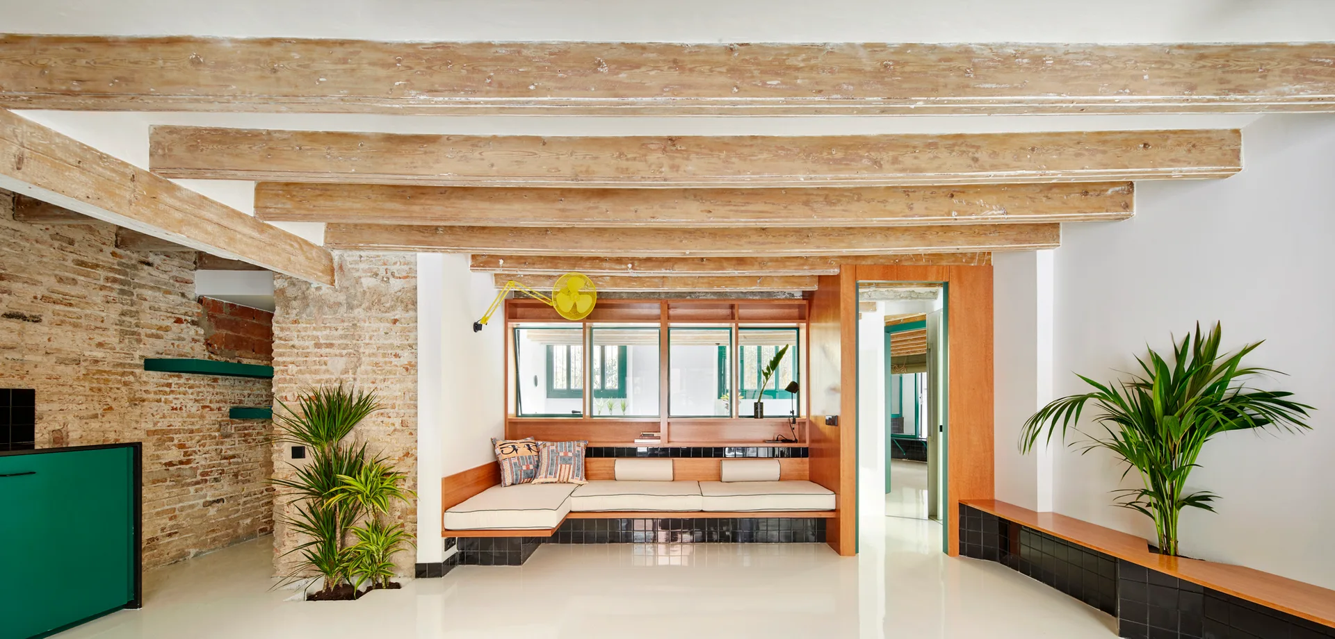A modern living room features a long built-in wooden bench with a black tiled base, exposed wooden ceiling beams, and glossy white floors.