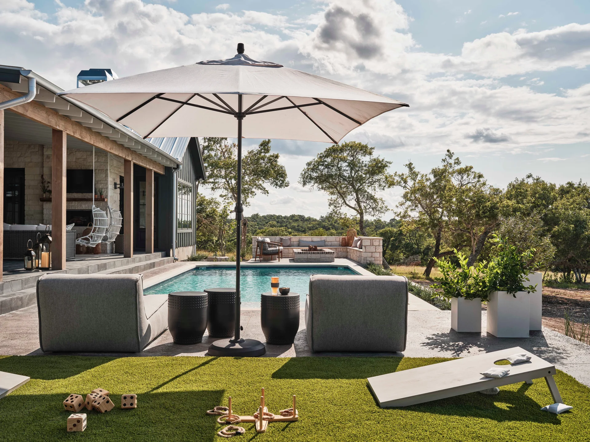 An outdoor swimming pool surrounded by a stone deck with gray lounge chairs, a large umbrella, and a green turf lawn with games.