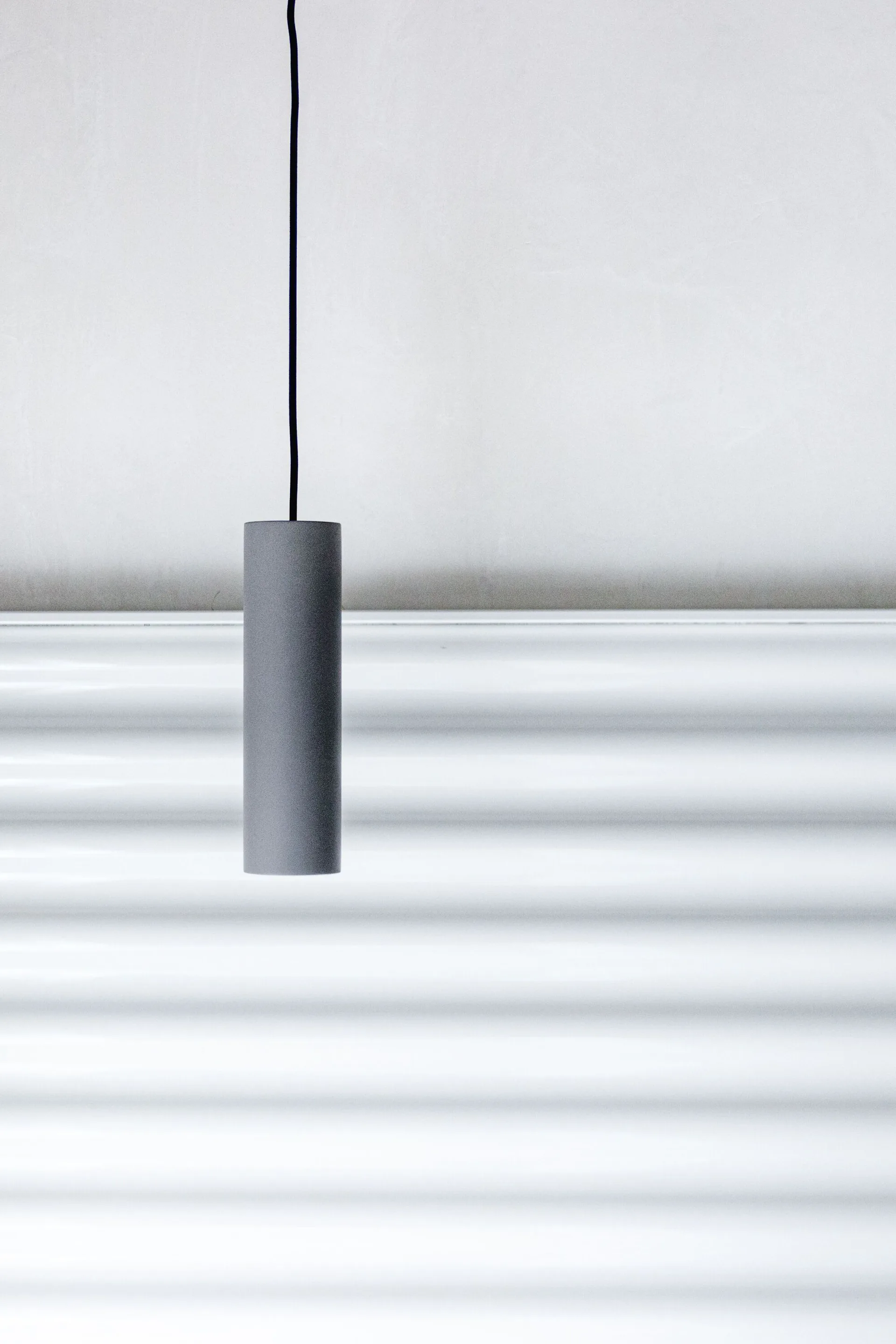 A matte gray cylindrical pendant light hangs from a black cord in front of a white wall with horizontal ribbed texture.