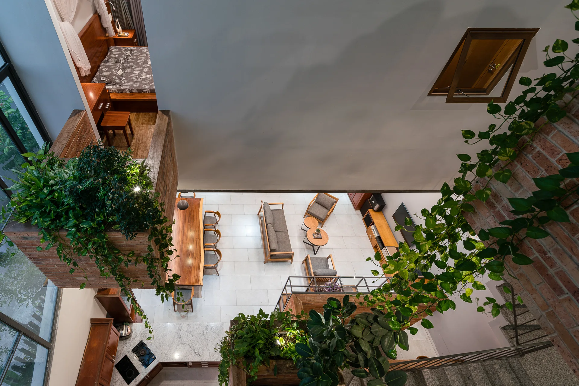 A high-angle view of a modern loft living room with grey seating, a wood dining table, exposed brick walls, and lush green hanging plants.