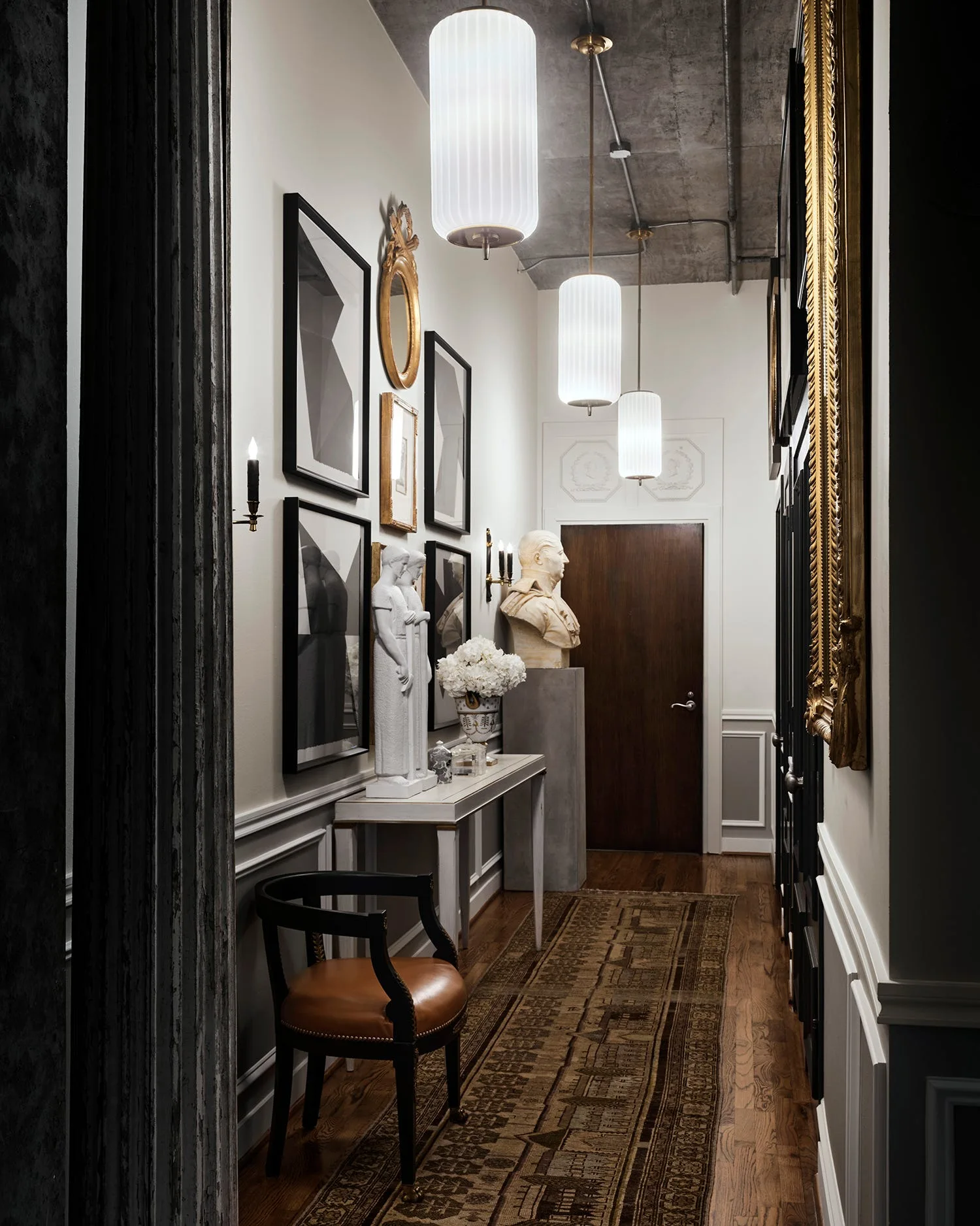 A narrow hallway featuring a raw concrete ceiling, three fluted glass pendant lights, paneled white walls with framed art, and a classical bust.