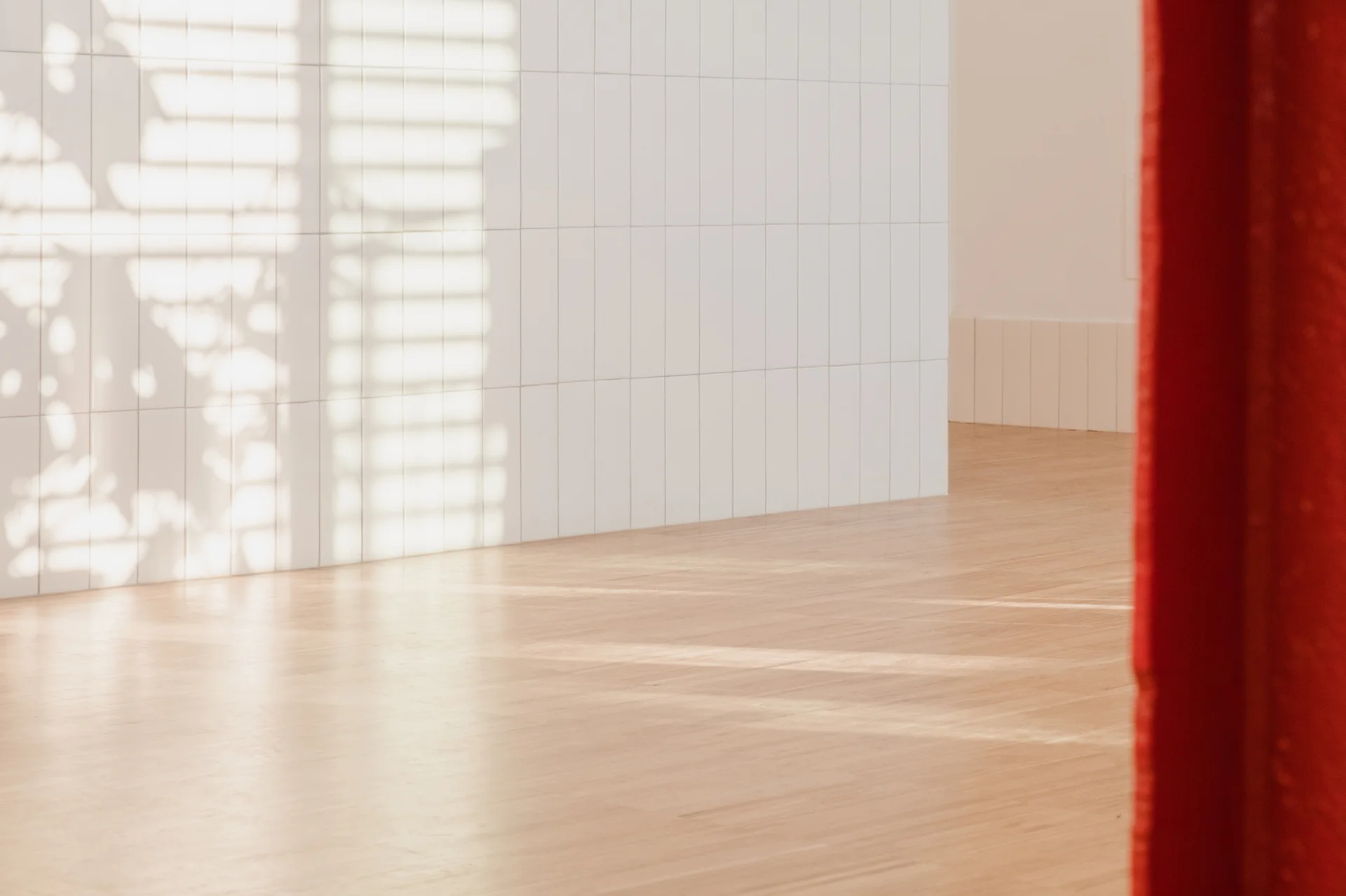 A wall of vertical white rectangular tiles meets light wood flooring in a bright space with leafy shadows and a red drape.