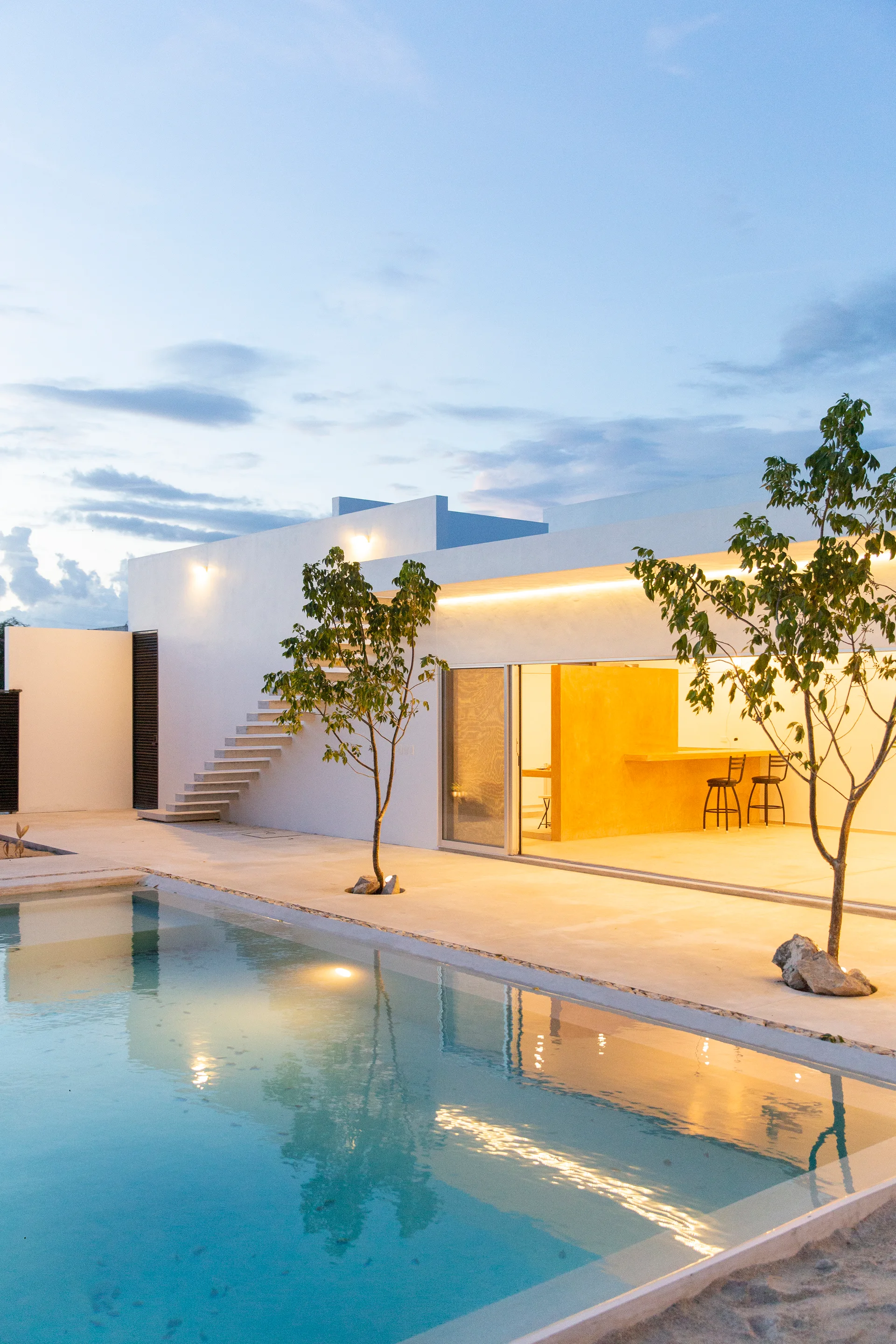 A modern white residential building features an outdoor pool, concrete patio, a floating staircase, and an illuminated yellow interior at dusk.