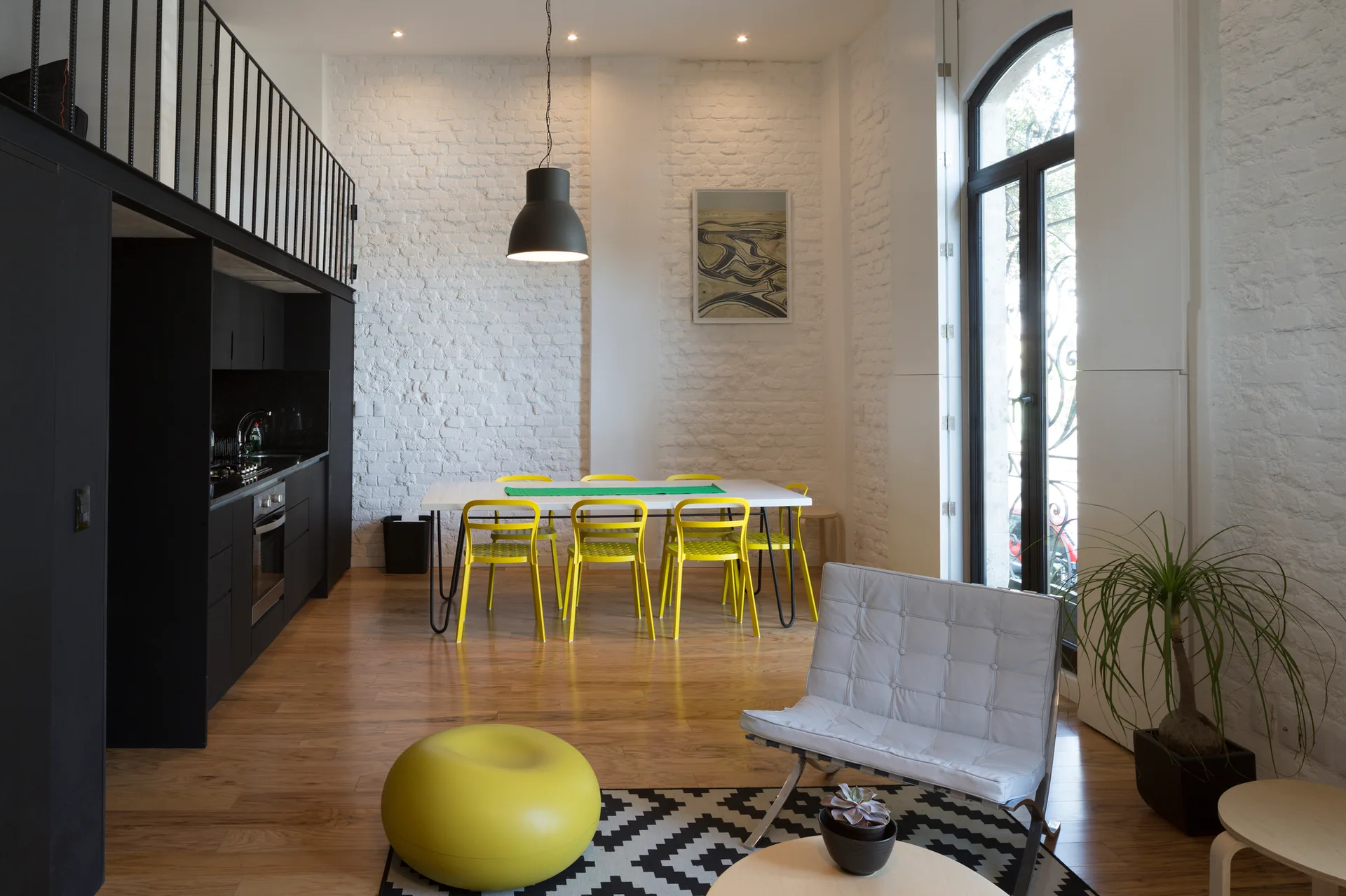 An open loft apartment featuring a white painted brick wall behind a dining table with yellow chairs and a black kitchen under a mezzanine.