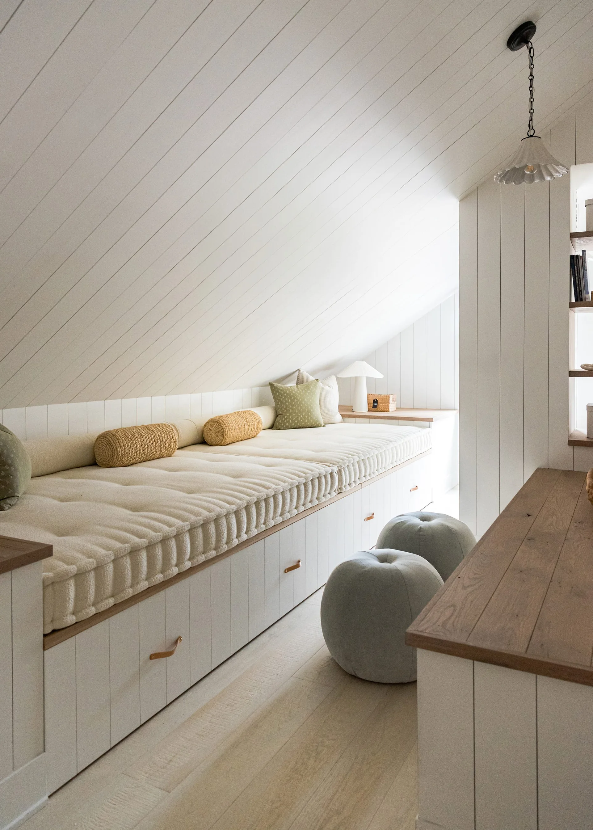 A custom built-in daybed with cream upholstery sits beneath a slanted shiplap ceiling in a cozy attic room next to two round grey ottomans.