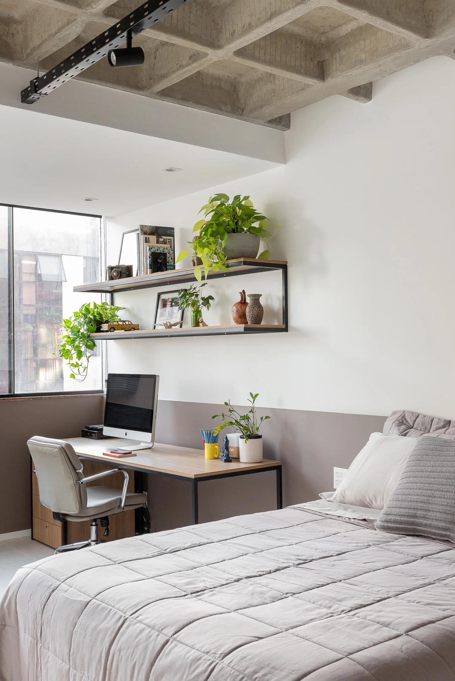 A bedroom with a gray quilted bed, a wood desk with a computer monitor, open wall shelves, and an exposed concrete waffle slab ceiling.