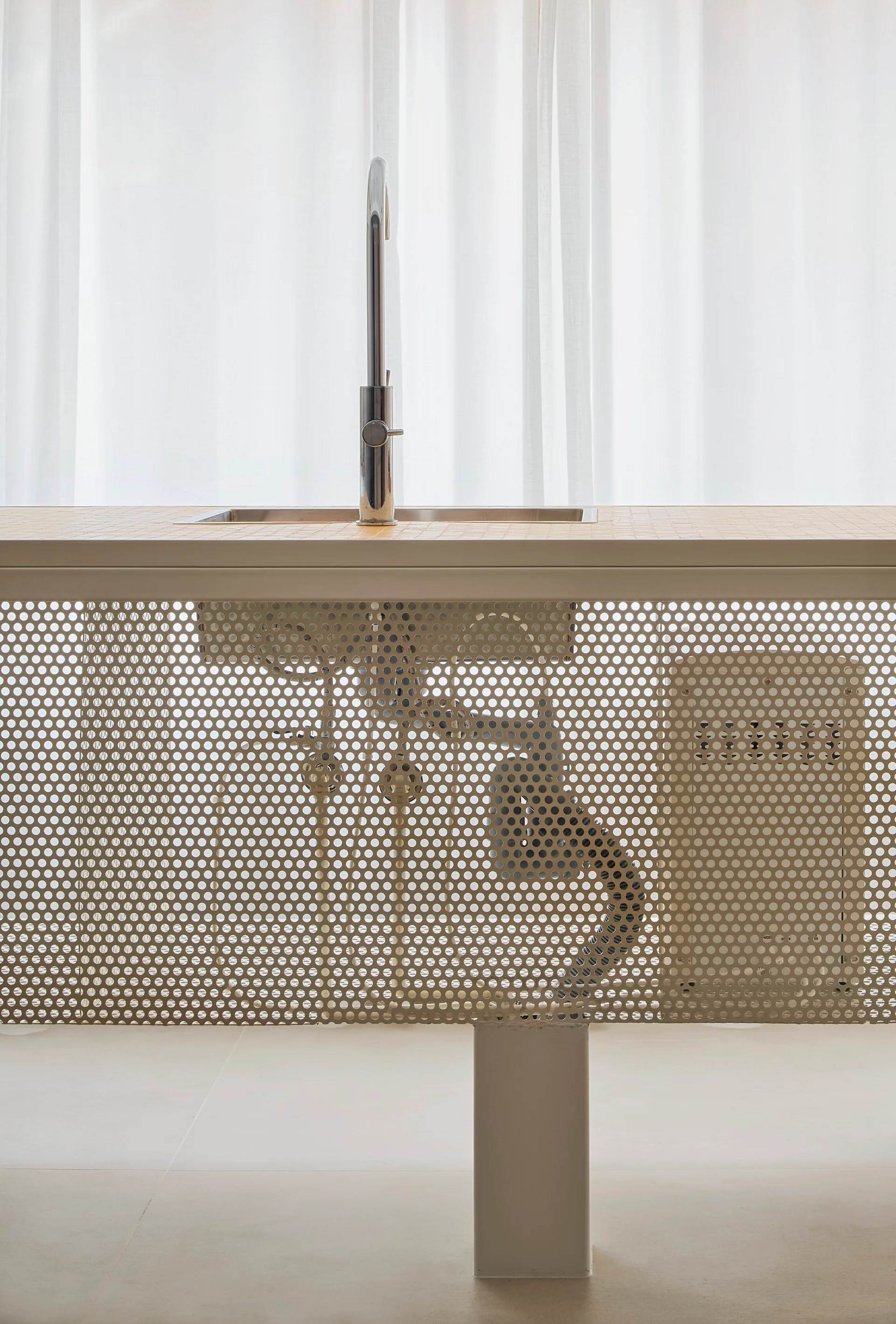 A minimalist kitchen island with a tiled countertop, chrome faucet, and a perforated cream metal screen showing under-sink plumbing against sheer curtains.