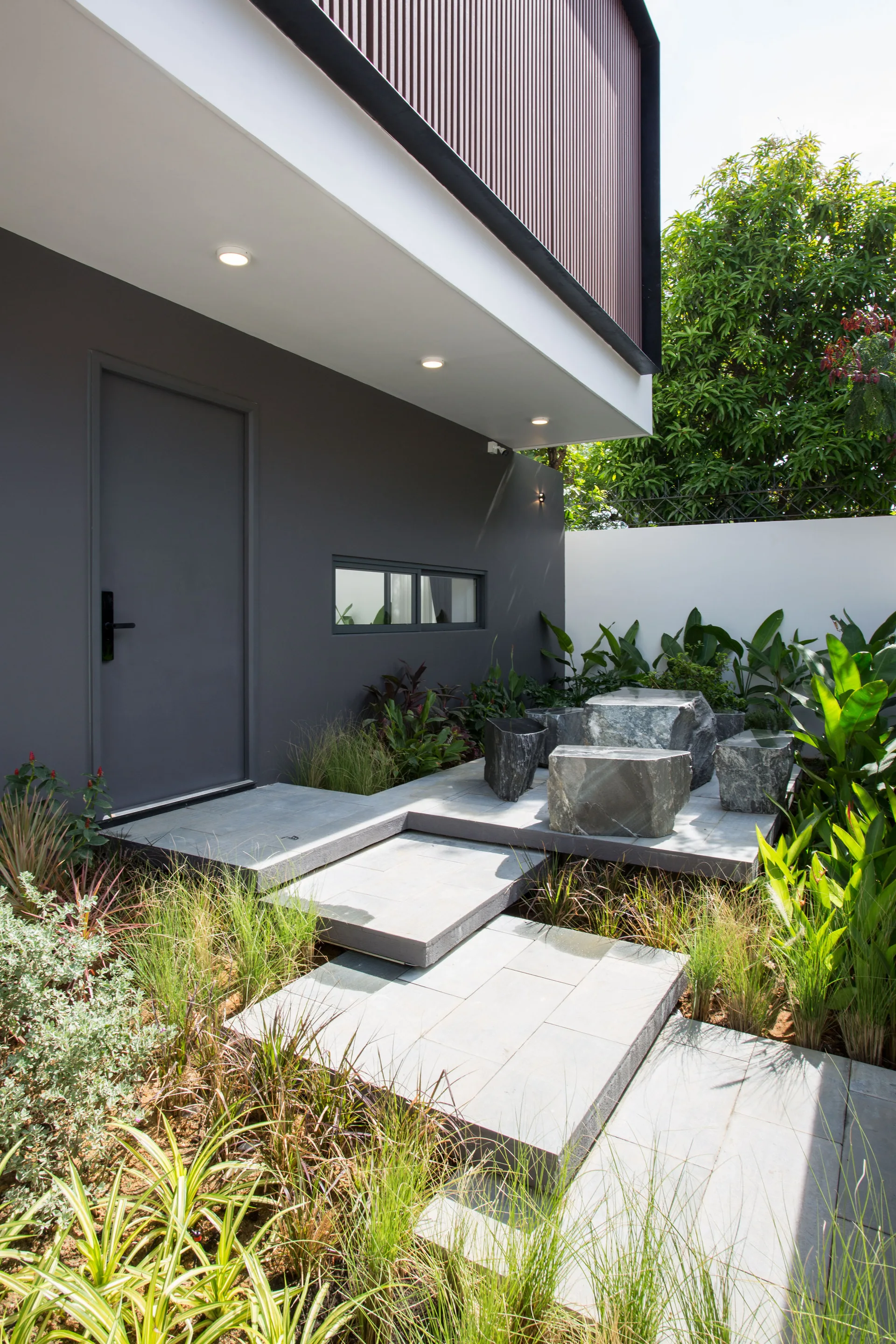 A modern outdoor entryway features stepped grey stone pavers, ornamental grasses, and raw stone blocks used as patio seating.