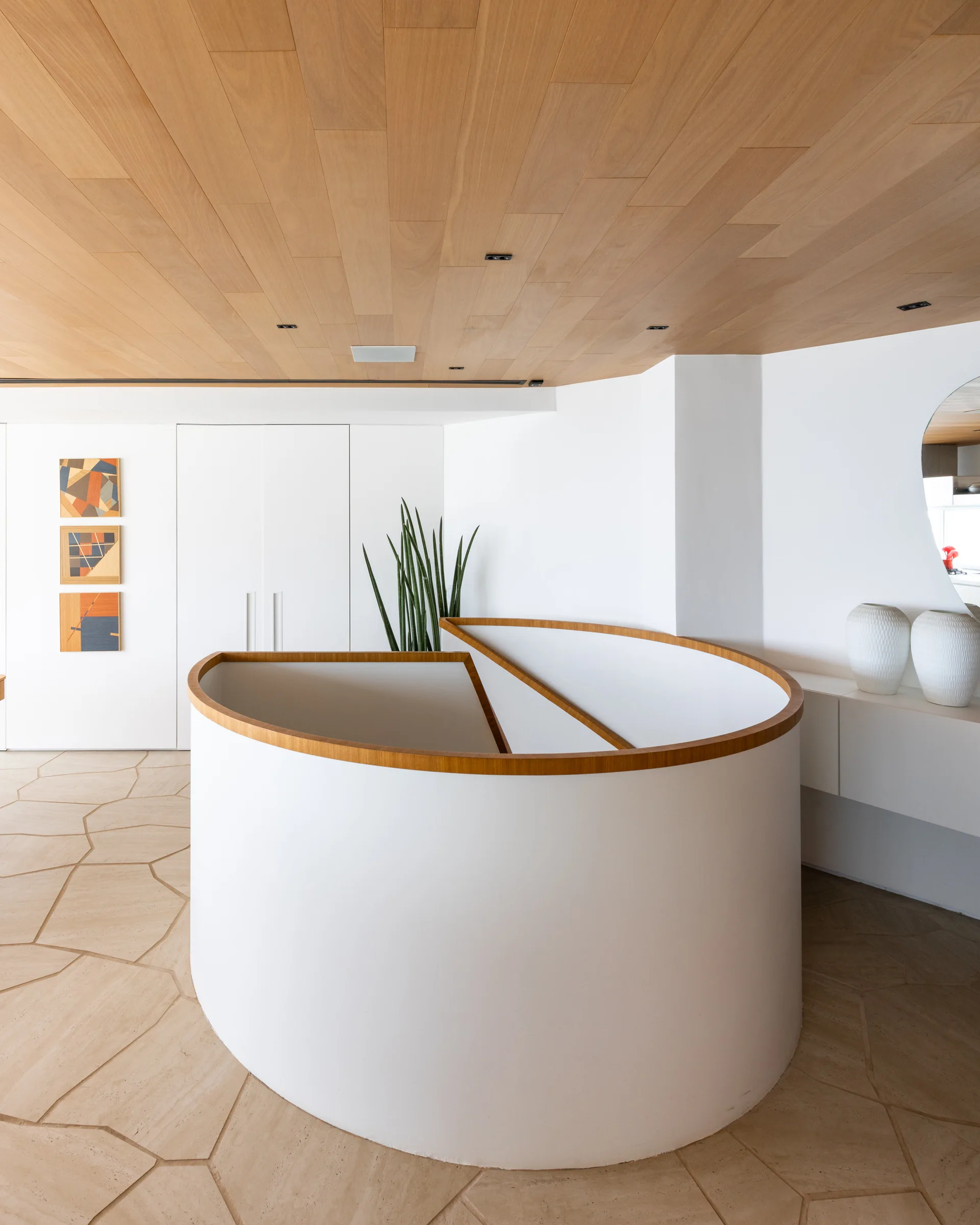 A curved white staircase with light wood trim sits beneath a wood plank ceiling in a minimalist space with irregular stone floor tiles.