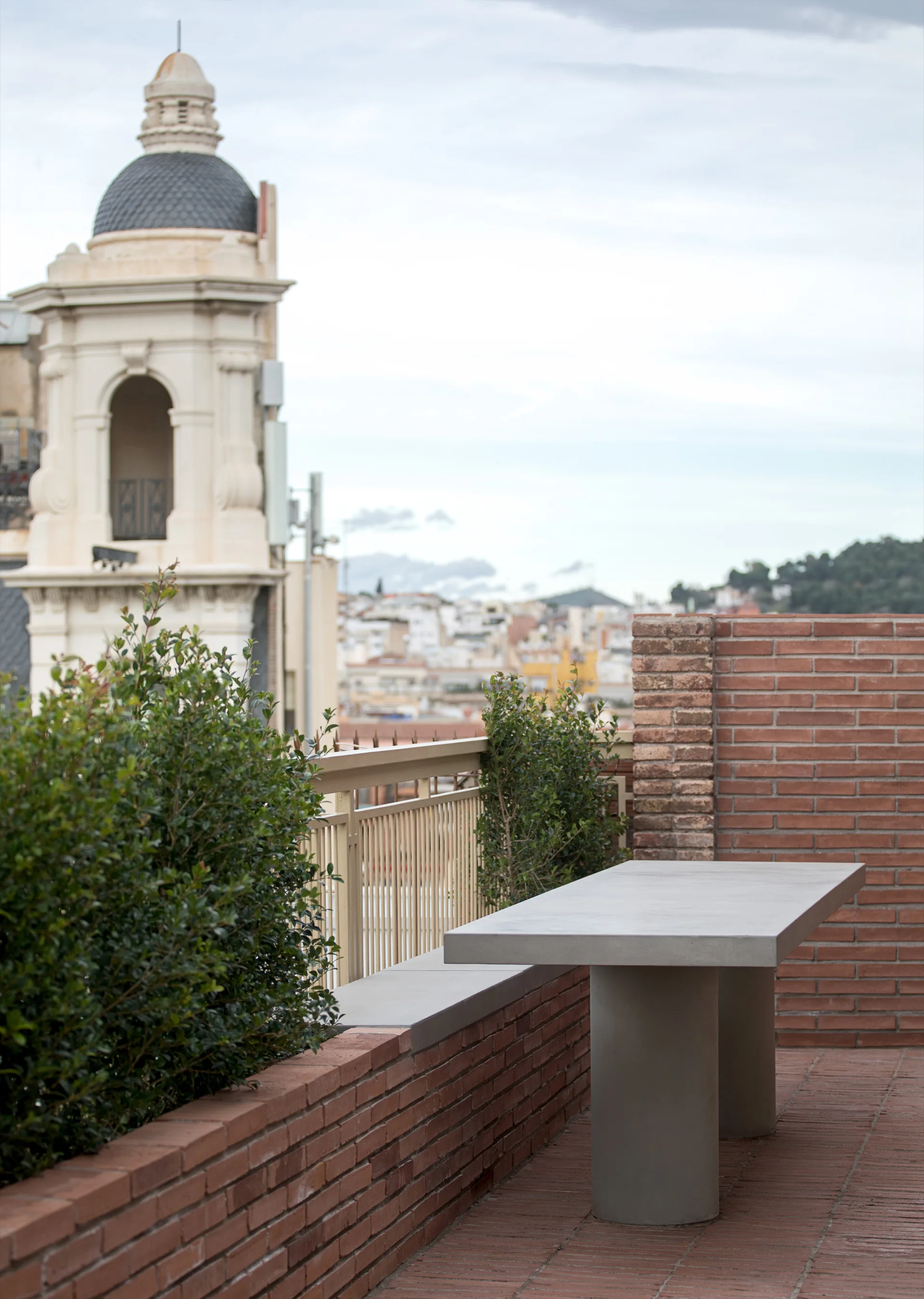 A minimalist concrete table stands on an outdoor rooftop terrace with a red brick wall, tiled flooring, and a city view in the background.