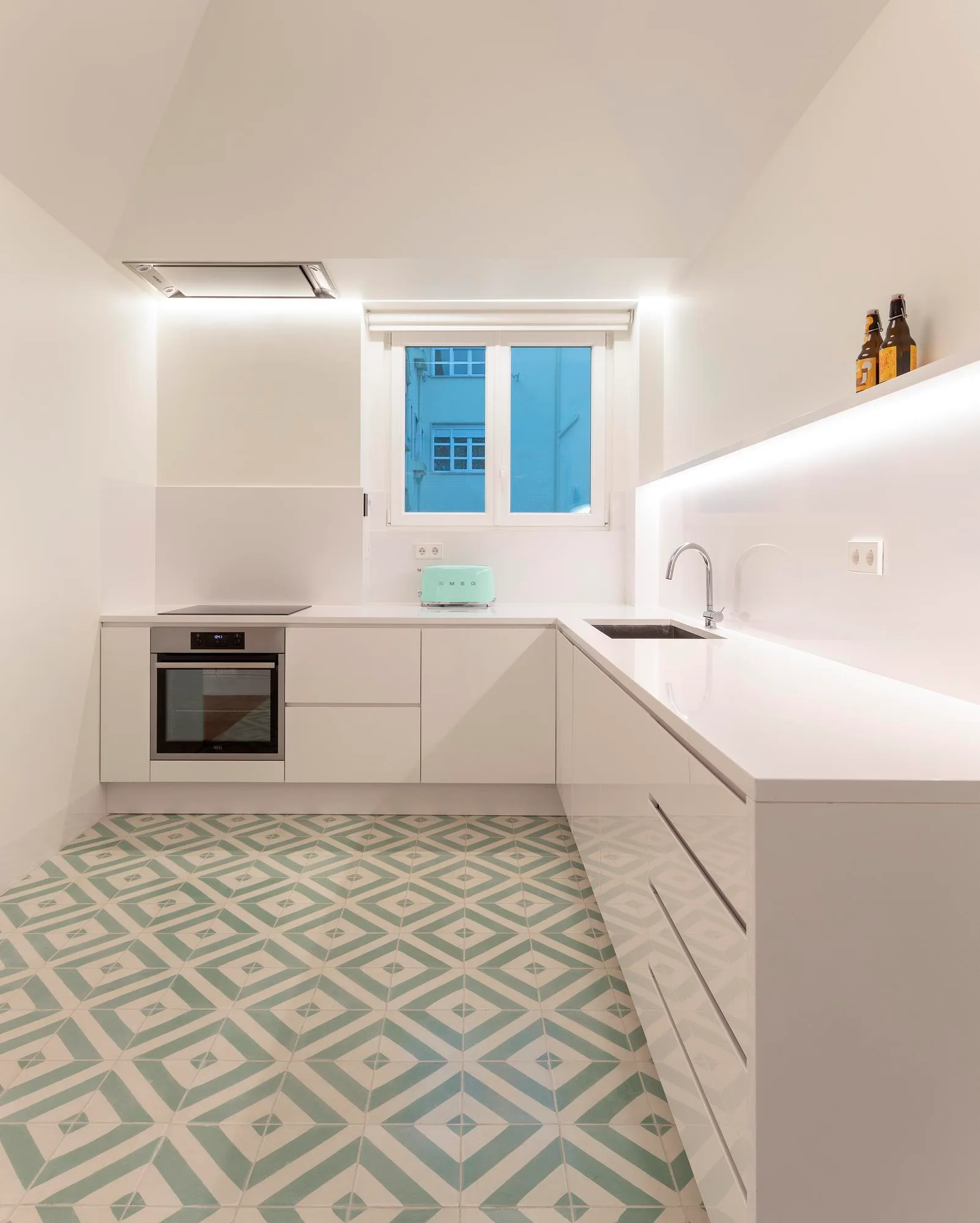 A minimalist white kitchen features flat-front cabinets, white countertops, a small window, and a green and white geometric tiled floor.