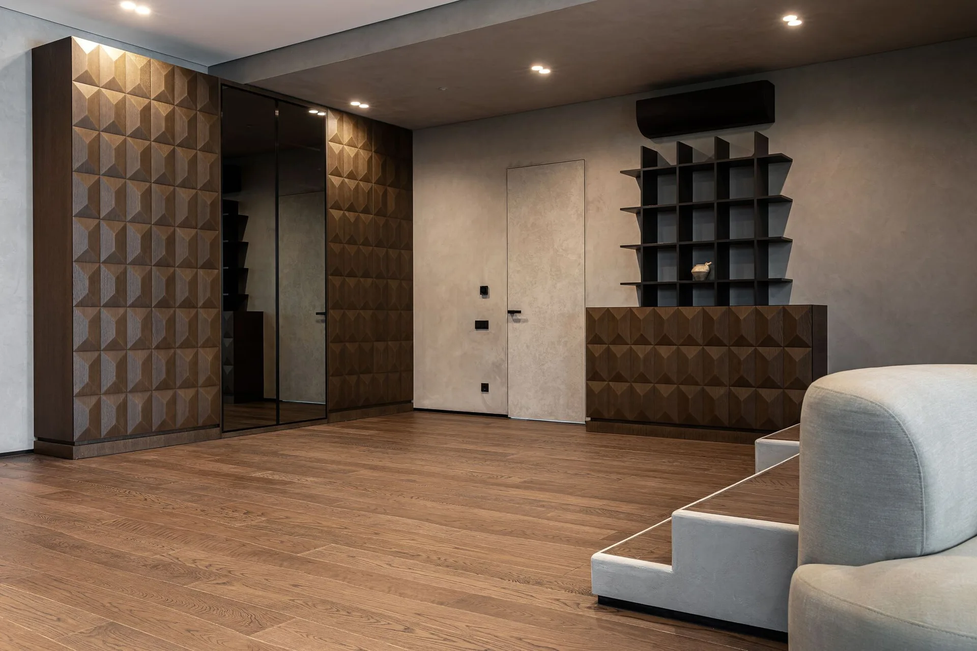 A modern living room features dark wood cabinetry with geometric pyramid patterns, a flush door in a grey plaster wall, and hardwood flooring.