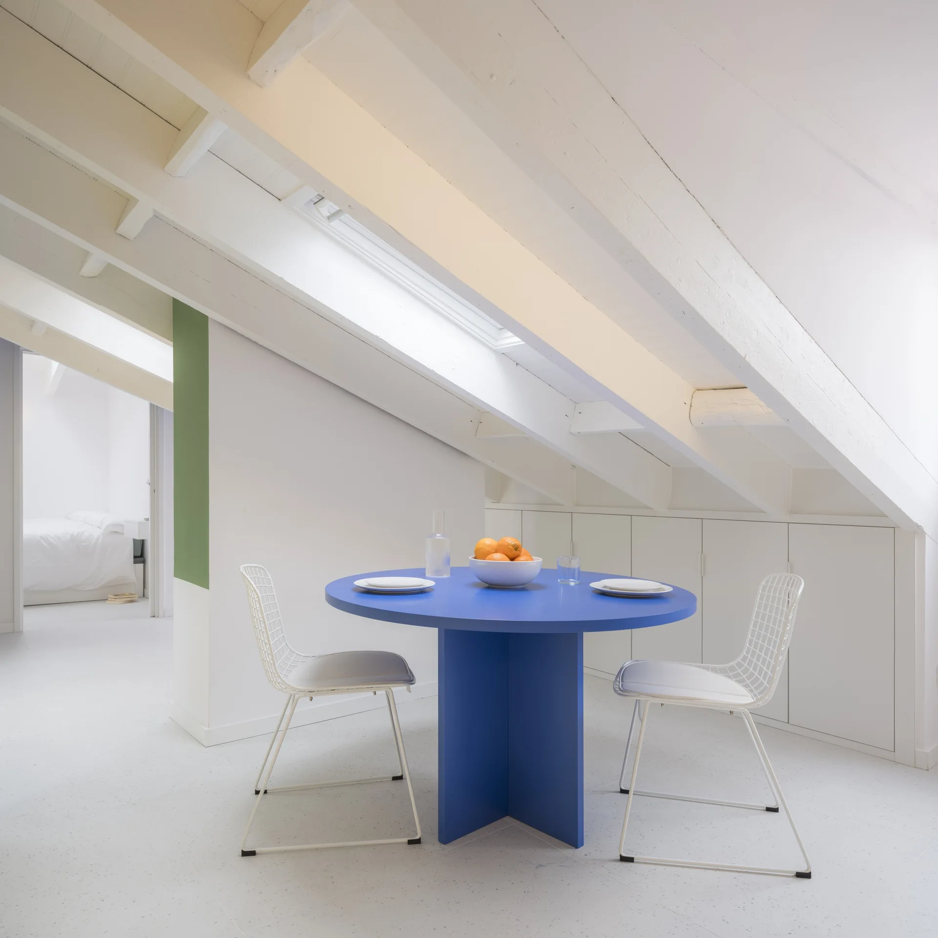 A bright attic dining space features a circular blue table and white wire chairs under a sloped ceiling with exposed beams and skylights.