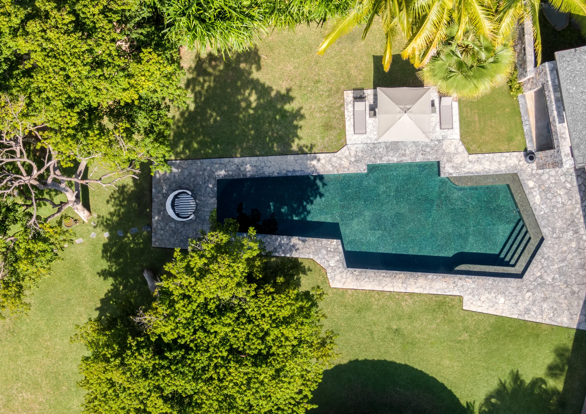 An aerial view of a dark green geometric swimming pool bordered by a gray flagstone deck, set within a lush grass lawn with mature trees.