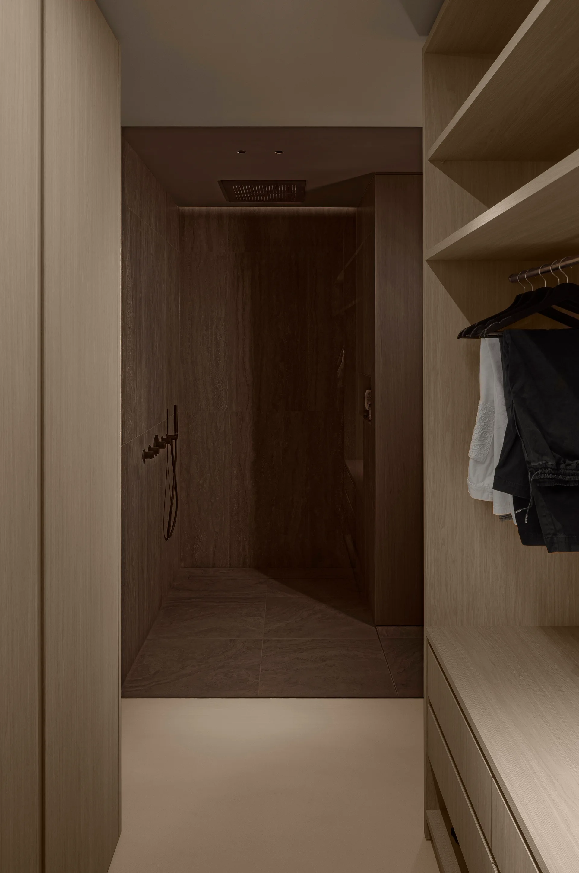 A view through a light wood dressing room wardrobe looking into a dark brown stone-tiled walk-in shower.