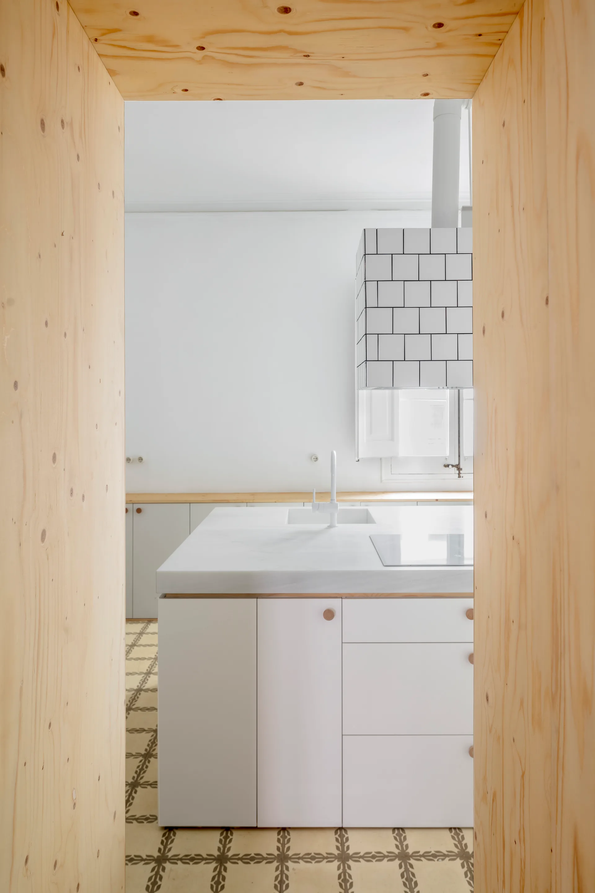 A minimalist white kitchen with a tiled range hood and patterned floor tiles, viewed through a natural plywood archway.