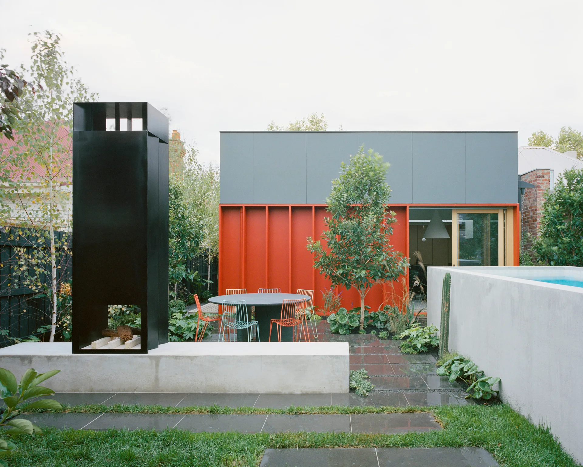 A modern backyard patio features a circular dining set, a tall black metal fireplace on a concrete base, and a vibrant orange exterior wall.
