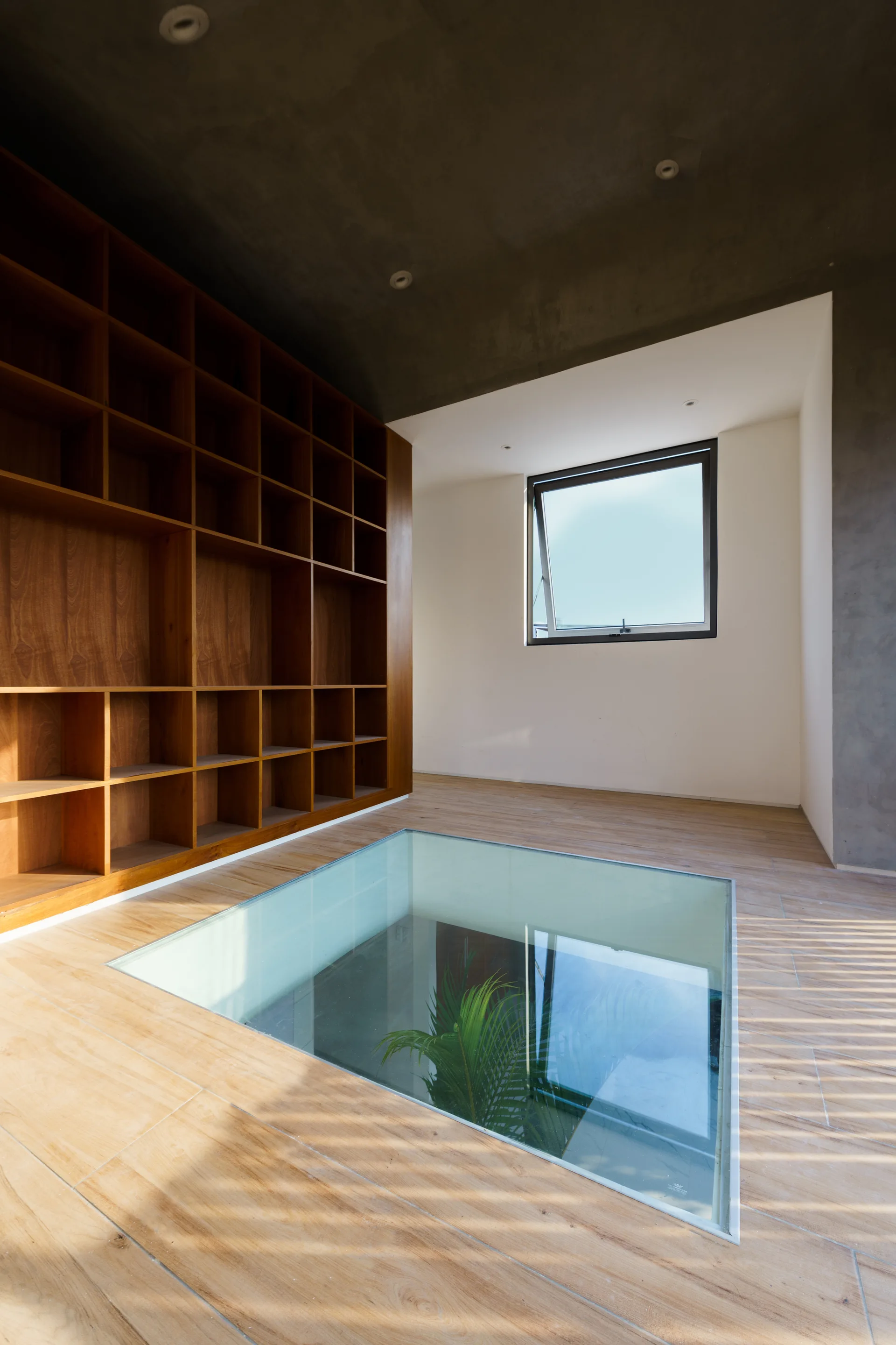 A modern room featuring a large wood grid bookshelf, a grey concrete-textured ceiling, and a transparent glass floor panel showing a plant below.