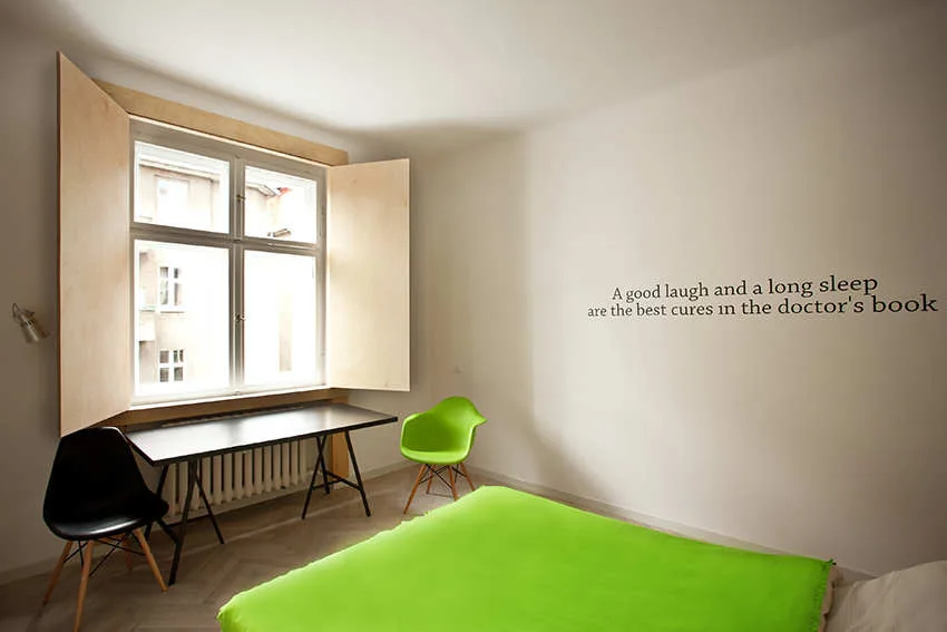 A minimalist bedroom featuring a bed with a bright green blanket, a black desk under a window with light wood shutters, and two molded plastic chairs.