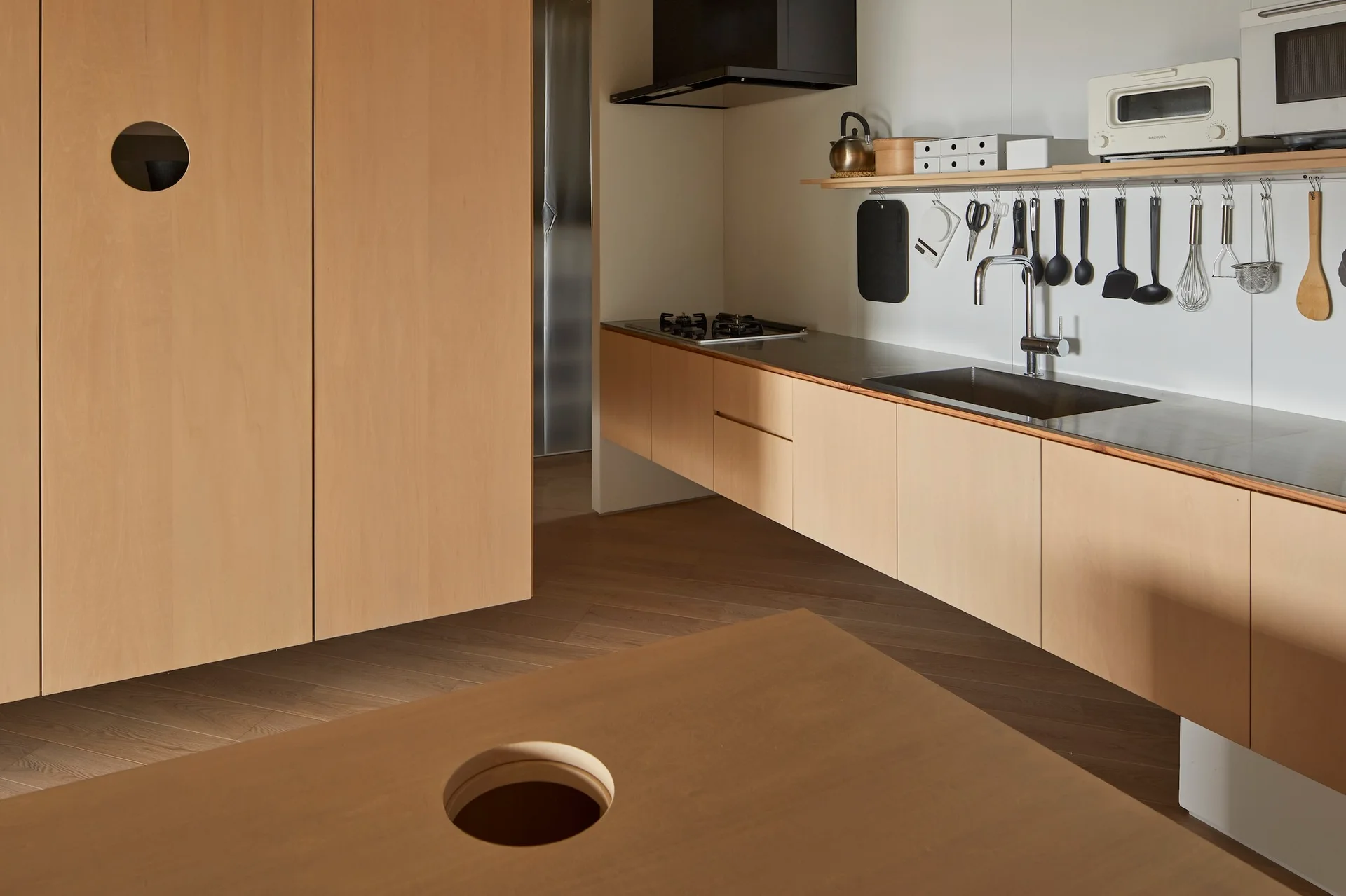 A minimalist kitchen features light wood cabinets, a stainless steel countertop with a sink, and a wall-mounted shelf with hanging utensils.