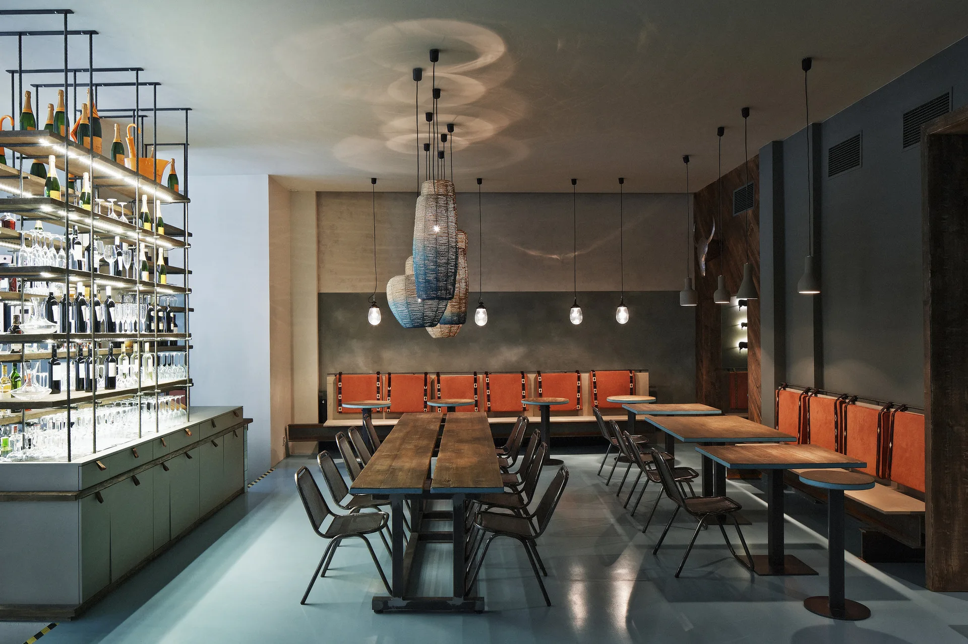 A modern restaurant dining room features long wooden tables, industrial metal chairs, orange cushioned banquette seating, and blue woven pendant lights.